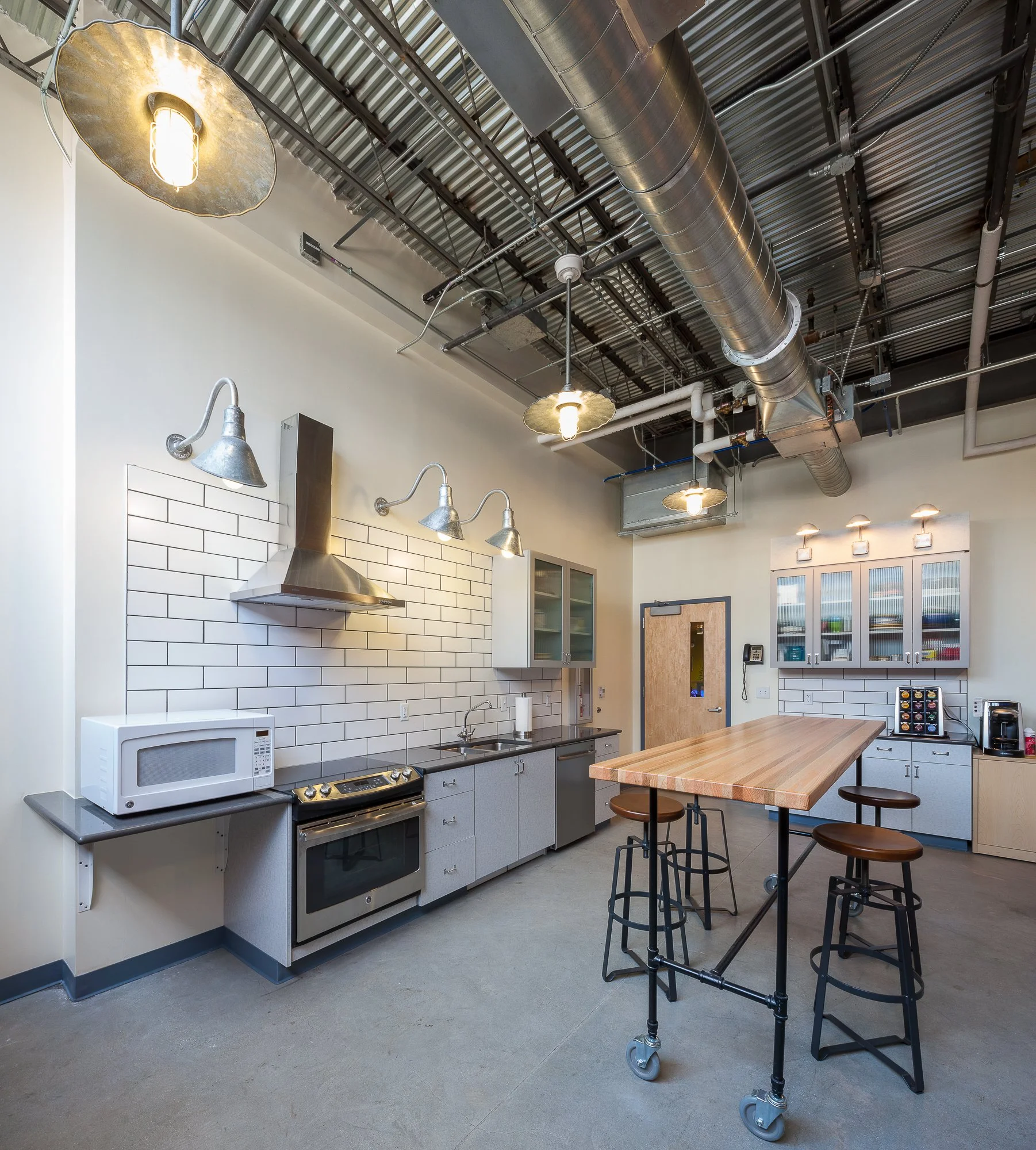 Industrial-style kitchen with white subway tile backsplash, gray cabinetry, and stainless steel appliances. A wooden island with black metal legs and casters is in the center, with three black metal stools. Overhead, there is exposed ductwork, pipes,