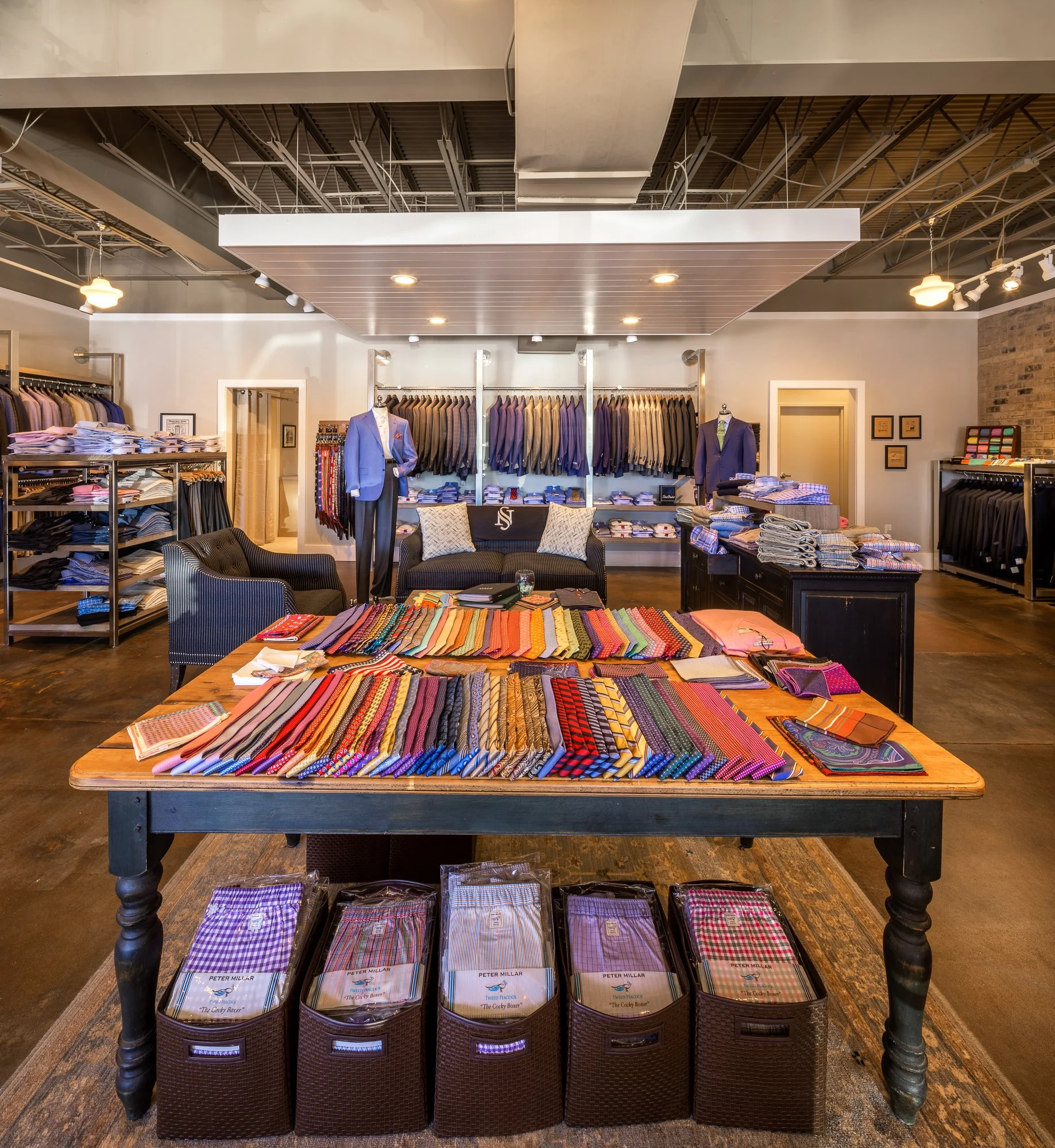 Indoor clothing store display featuring shelves of folded fabrics and racks of suits and shirts, with mannequins dressed in suits and a table of colorful neckties.
