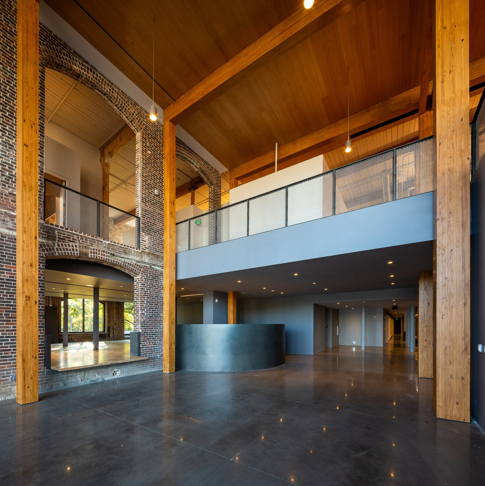 Interior of a modern building with wooden beams, exposed brick arches, hanging lights, and a balcony with a metal railing.
