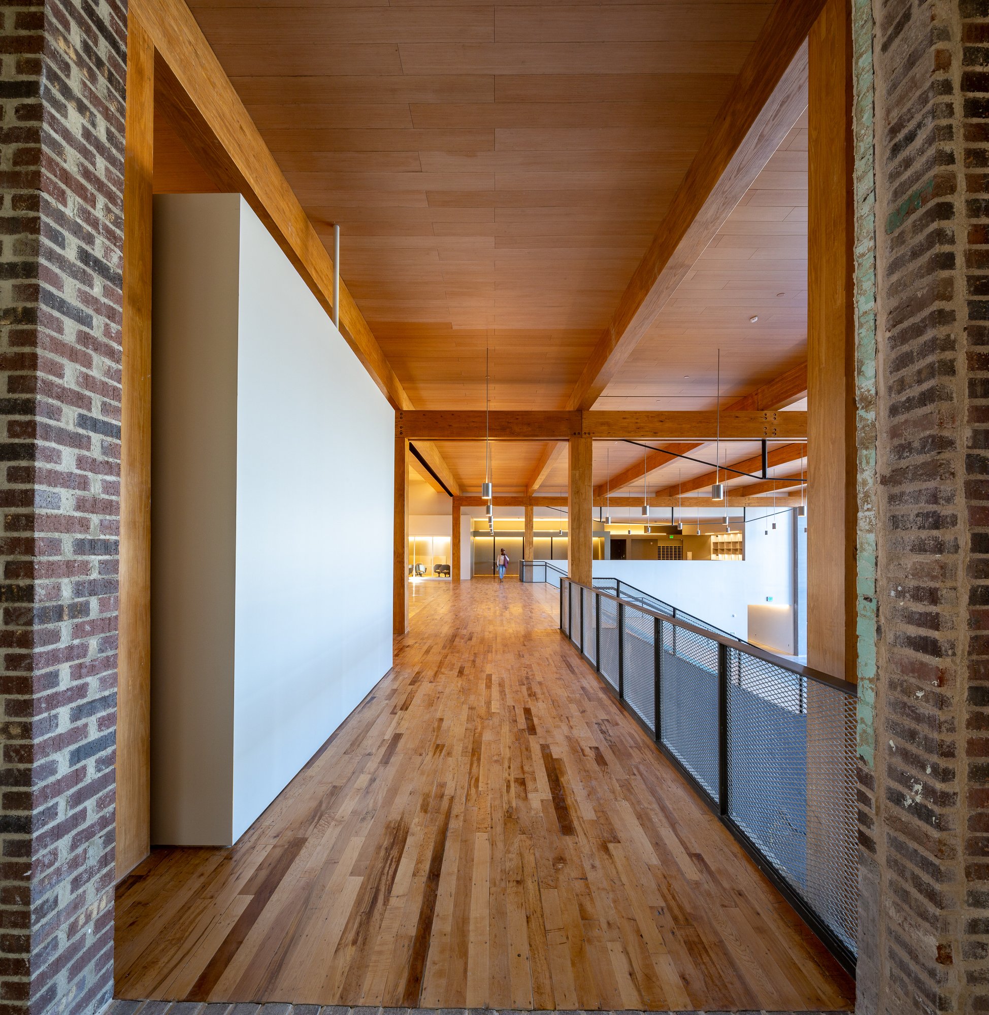 Interior view of a modern building with a wooden ceiling and floor, brick walls, and hanging pendant lights, featuring a person walking in the background.