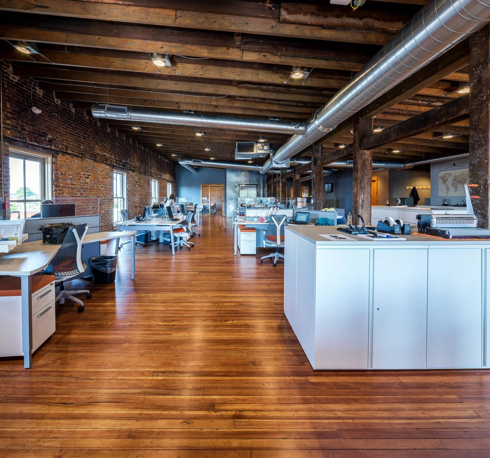 Open office space with wooden floors, exposed brick walls, high ceiling with wooden beams, visible air ducts, and multiple workstations with computers and chairs.