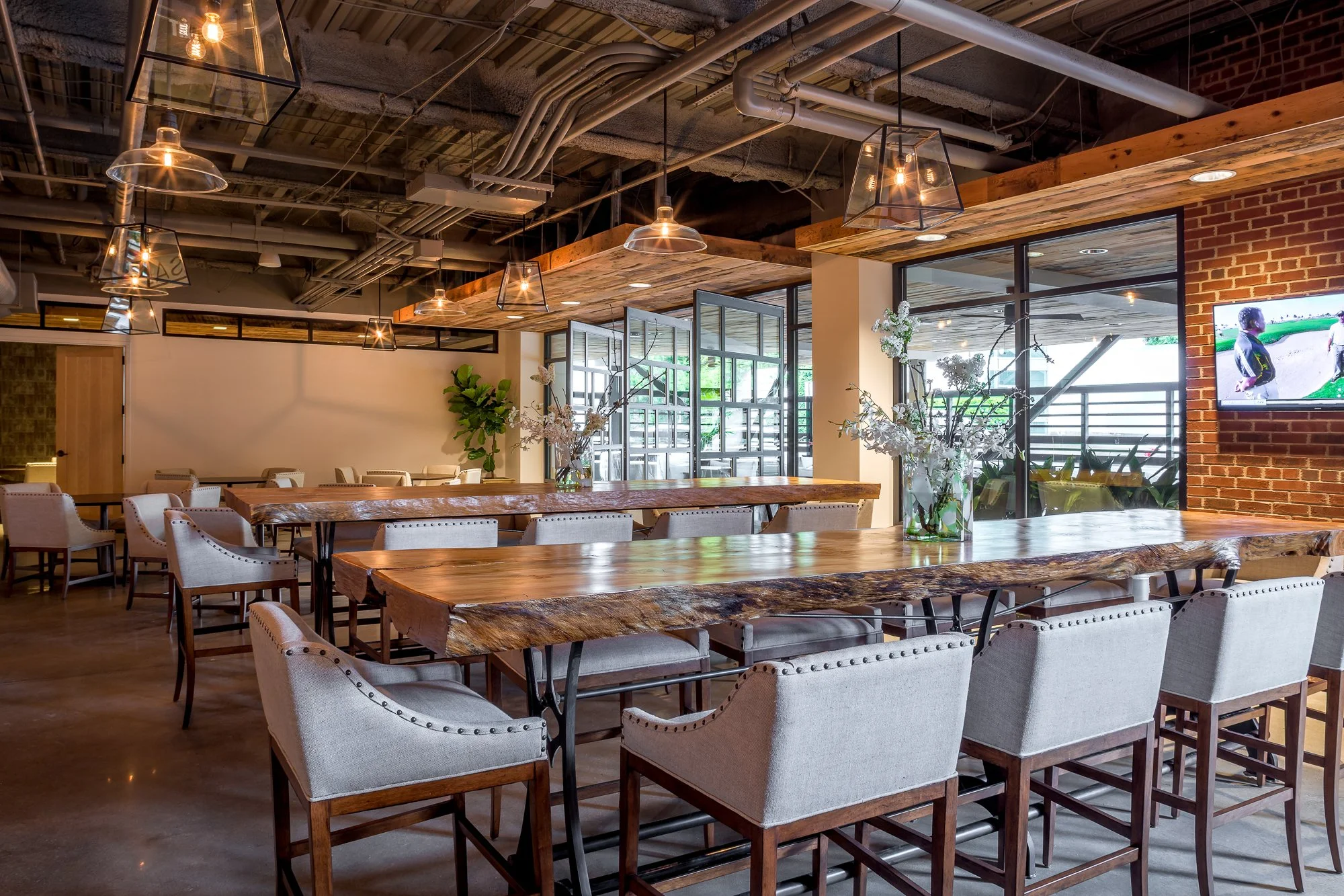 A modern restaurant interior featuring wooden tables, beige upholstered chairs with nailhead trim, and a brick wall with a mounted flat-screen TV. The ceiling has exposed pipes and wooden accents, and large windows let in natural light.