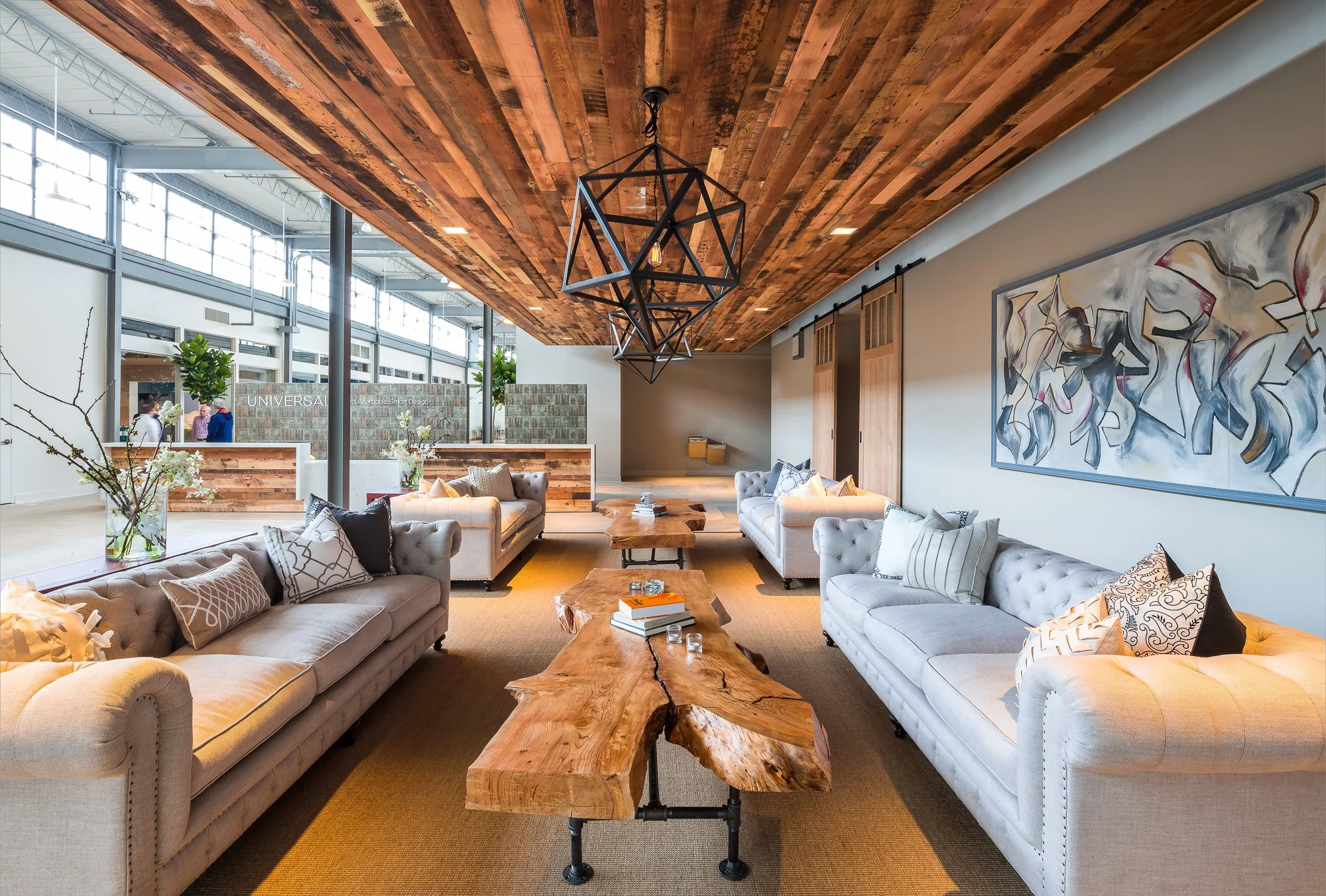 Modern lounge area with two large white sofas adorned with decorative pillows, a rustic wooden coffee table with books and candles, a contemporary artwork on the wall, and a striking geometric chandelier hanging from a wood-paneled ceiling.