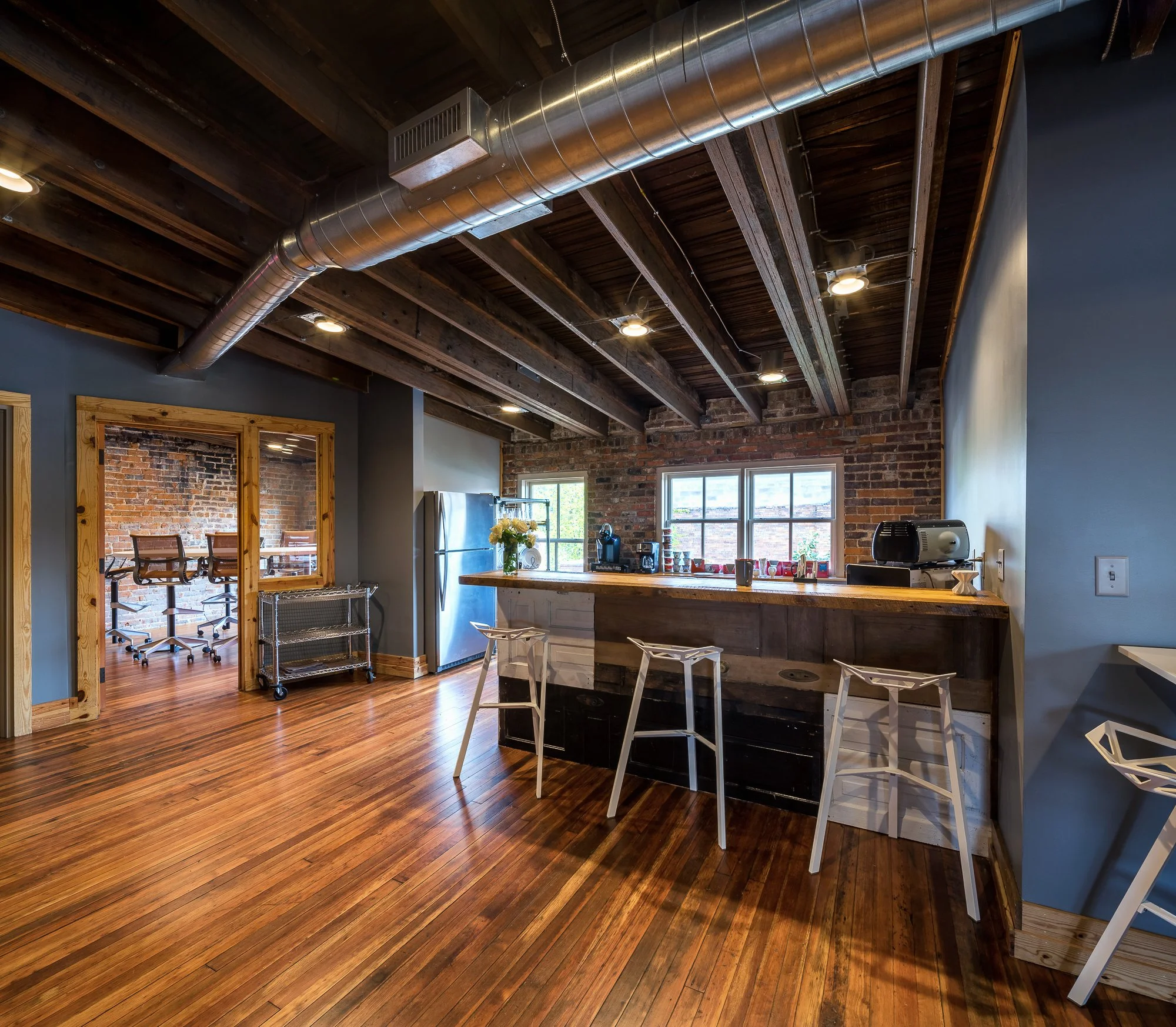 Interior view of a kitchen with exposed brick wall, wooden ceiling with metal ductwork, a window, and a dark kitchen counter with white chairs.