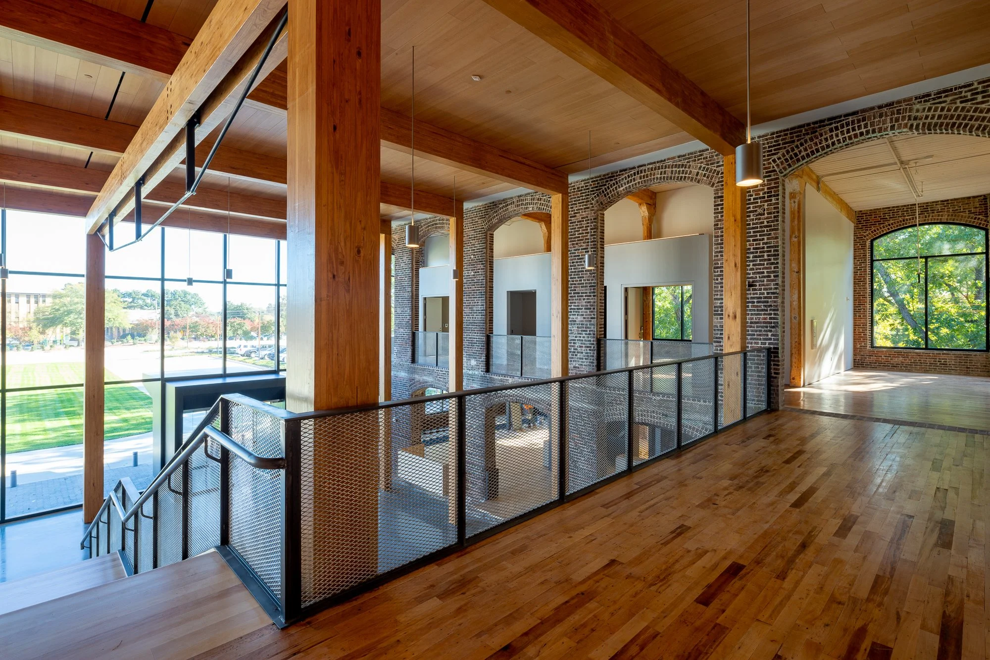 Interior of a modern building with large glass windows, wooden ceiling beams, brick walls, and a staircase with metal railings.