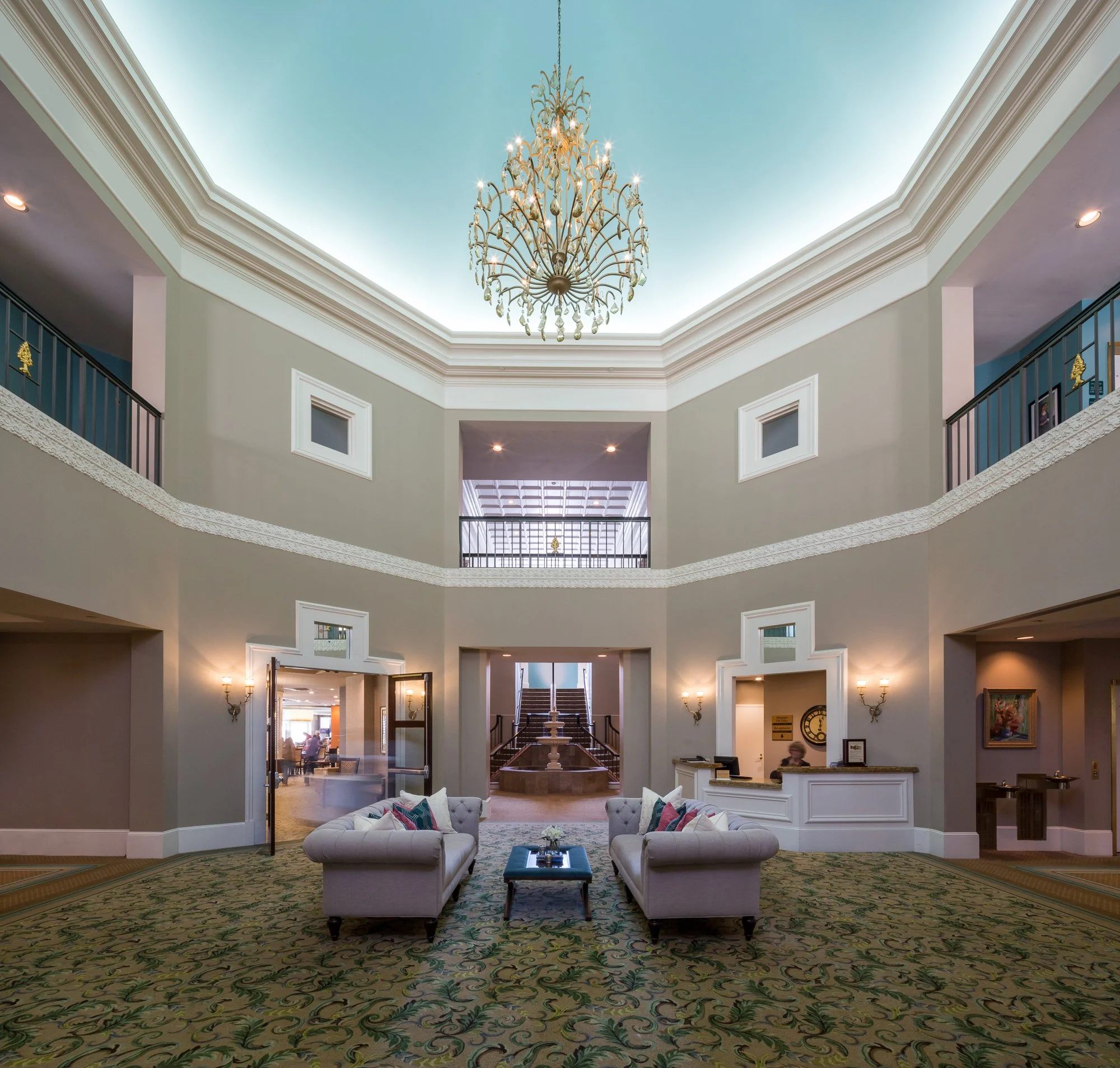 Hotel lobby with a green floral carpet, two white tufted sofas, a small table, and a large chandelier hanging from the ceiling. The space has an open layout with stairs at the back and a balcony railing on the sides.