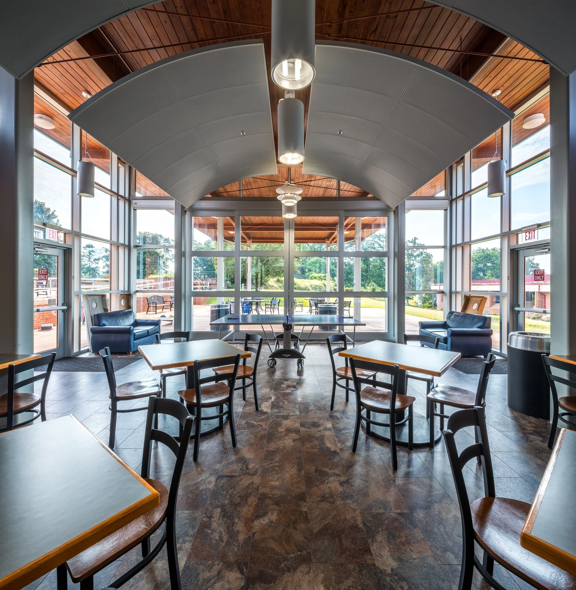 Interior of a modern building with a curved ceiling, large windows, and seating area with tables and chairs. Lounge chairs and patio outside are visible.