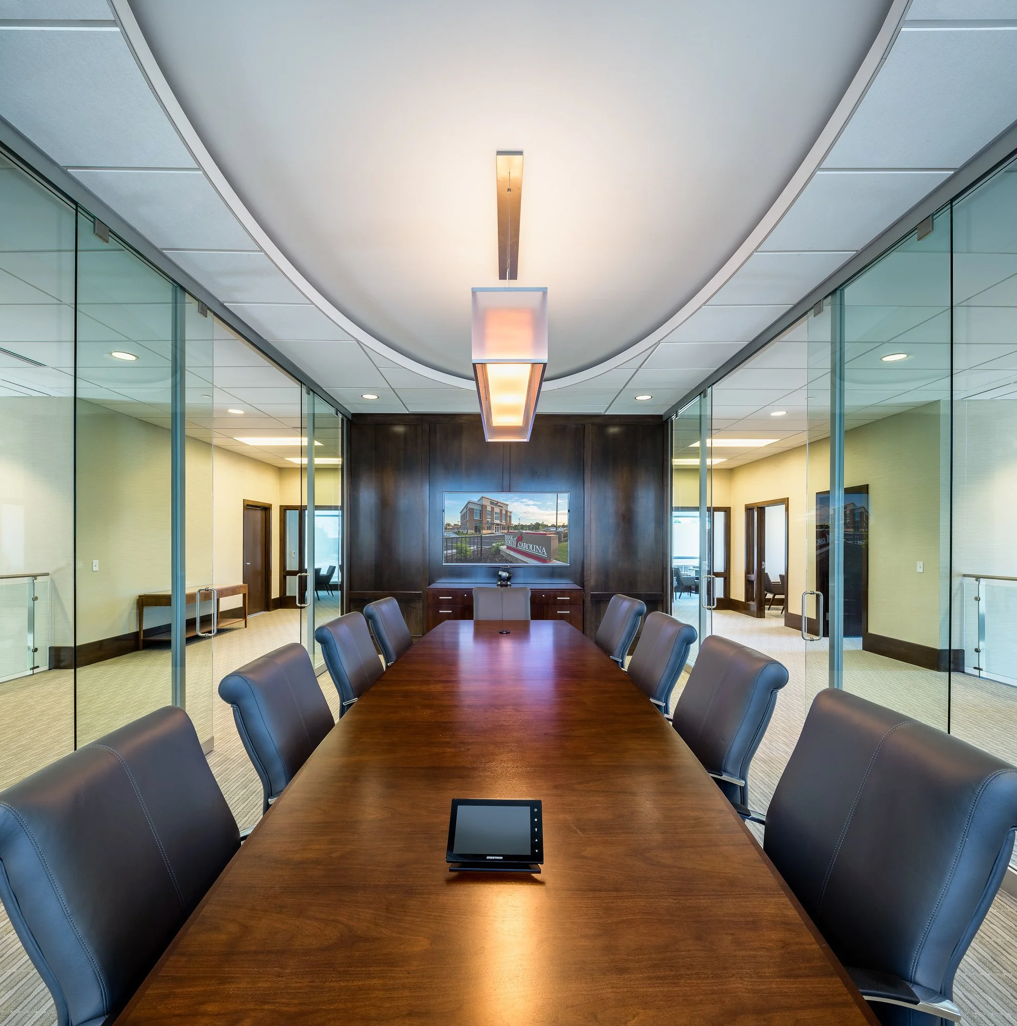 A modern conference room with a long wooden table surrounded by black leather chairs. There is a tablet on the table and a chandelier hanging from the ceiling. Glass walls reveal an office lobby outside, with a building and signs reading 'Bank of Nor