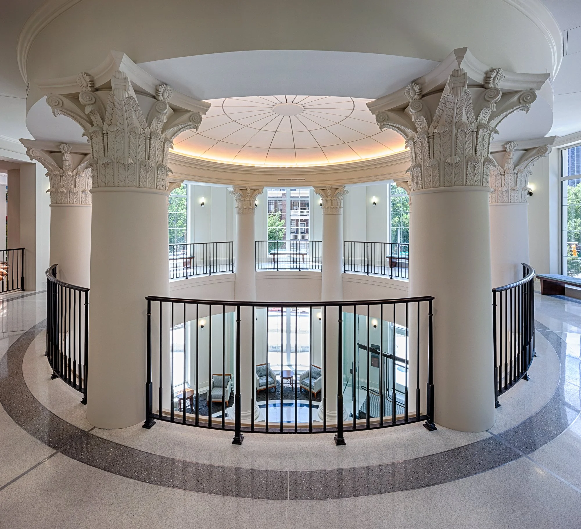 Interior view of a building with large round columns featuring ornate capitals, a circular balcony with a black railing, and large windows allowing natural light to illuminate the space