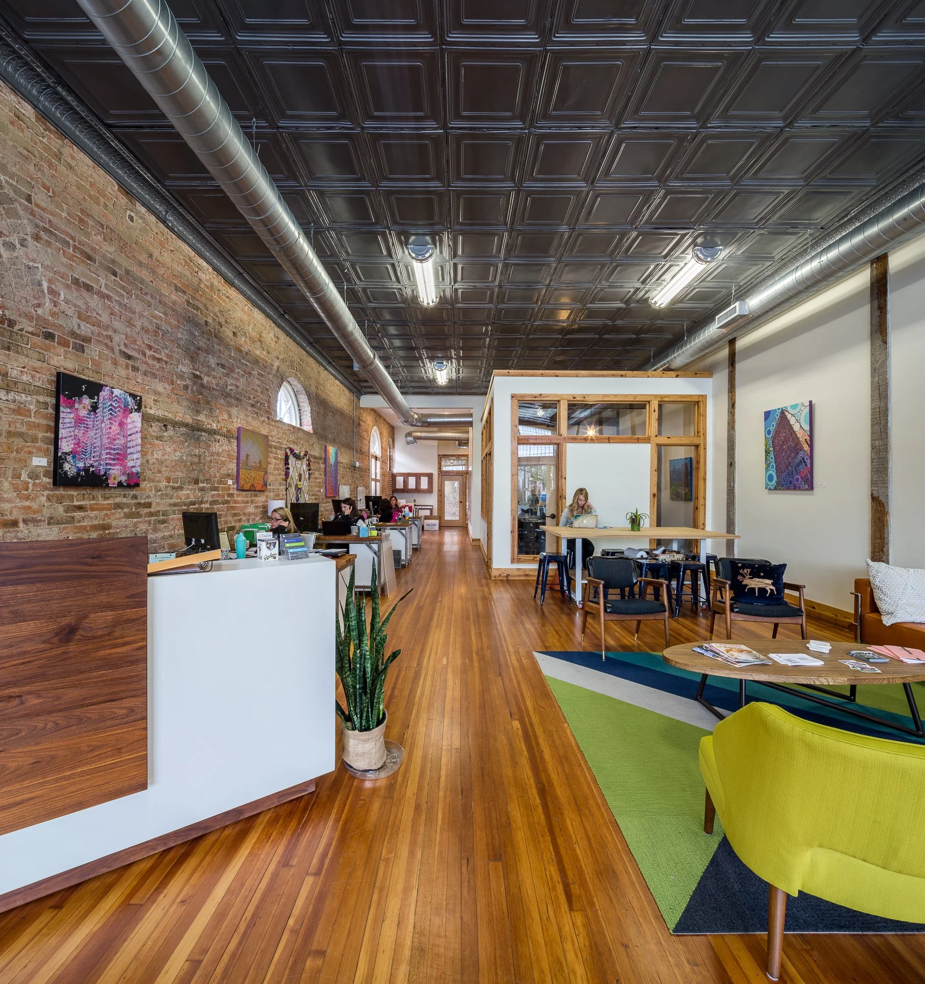 Interior of a modern office or coworking space with brick and white walls, colorful contemporary artwork, wooden furnishings, exposed ductwork, and a patterned rug. People are working at desks and tables.