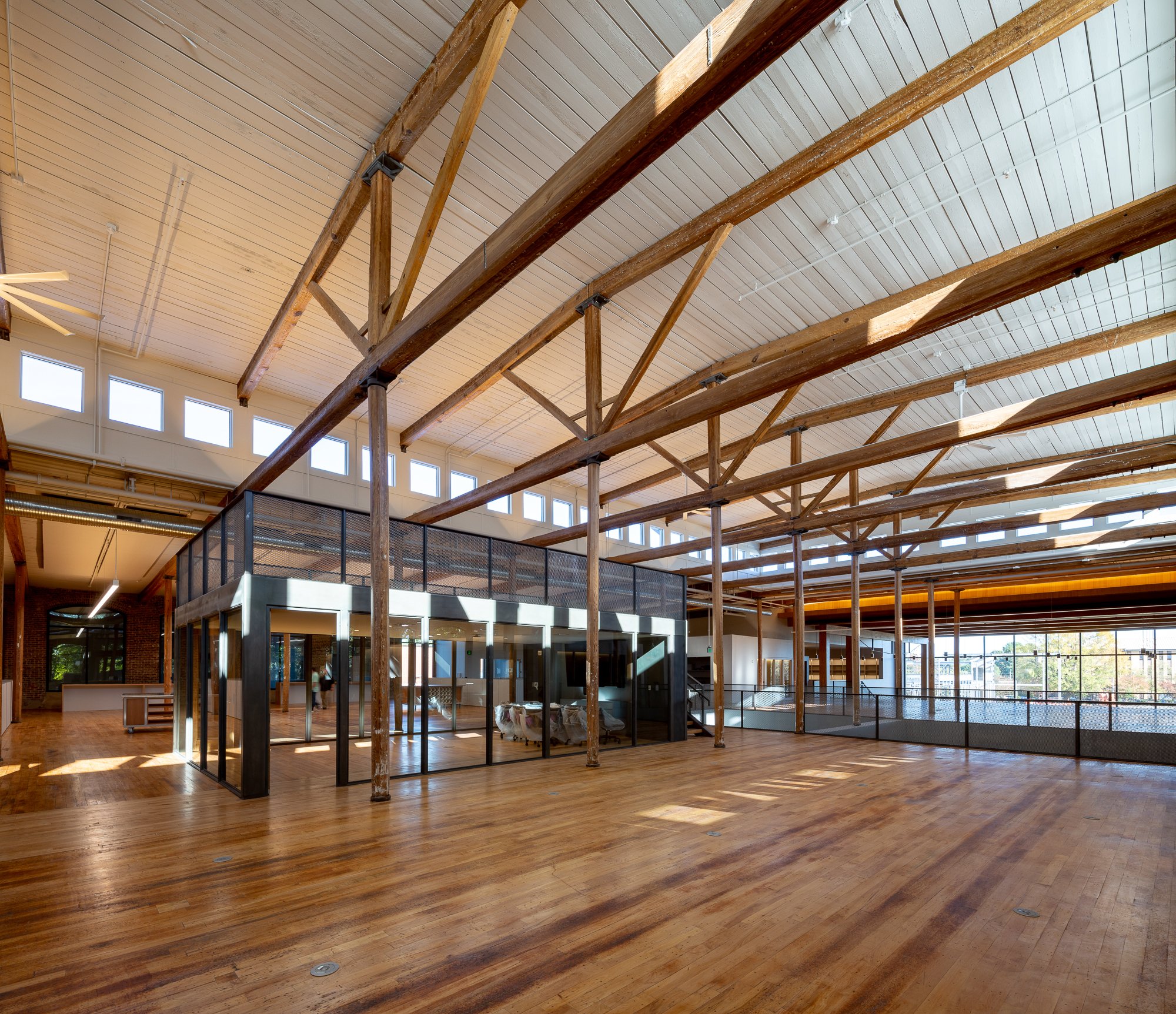 Interior of a building with wooden beams, a high ceiling, large windows, and a glass enclosed area at the center. The wooden floor is shiny and polished.
