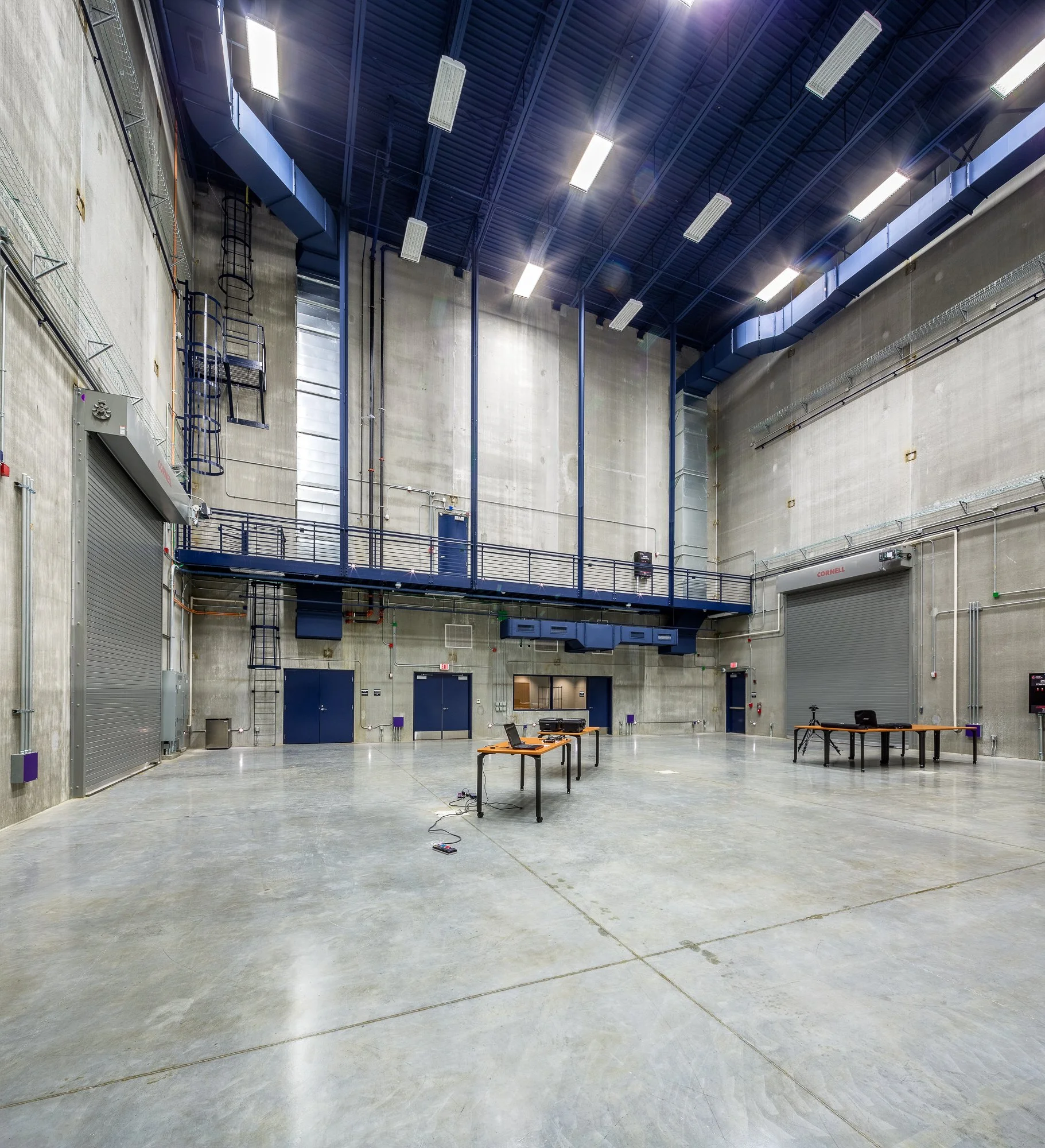 A large industrial or warehouse space with high ceilings and bright fluorescent lighting. There are tables with electronic equipment, chairs, and a tripod in the area. The walls are concrete, with various pipes and an overhead railing.