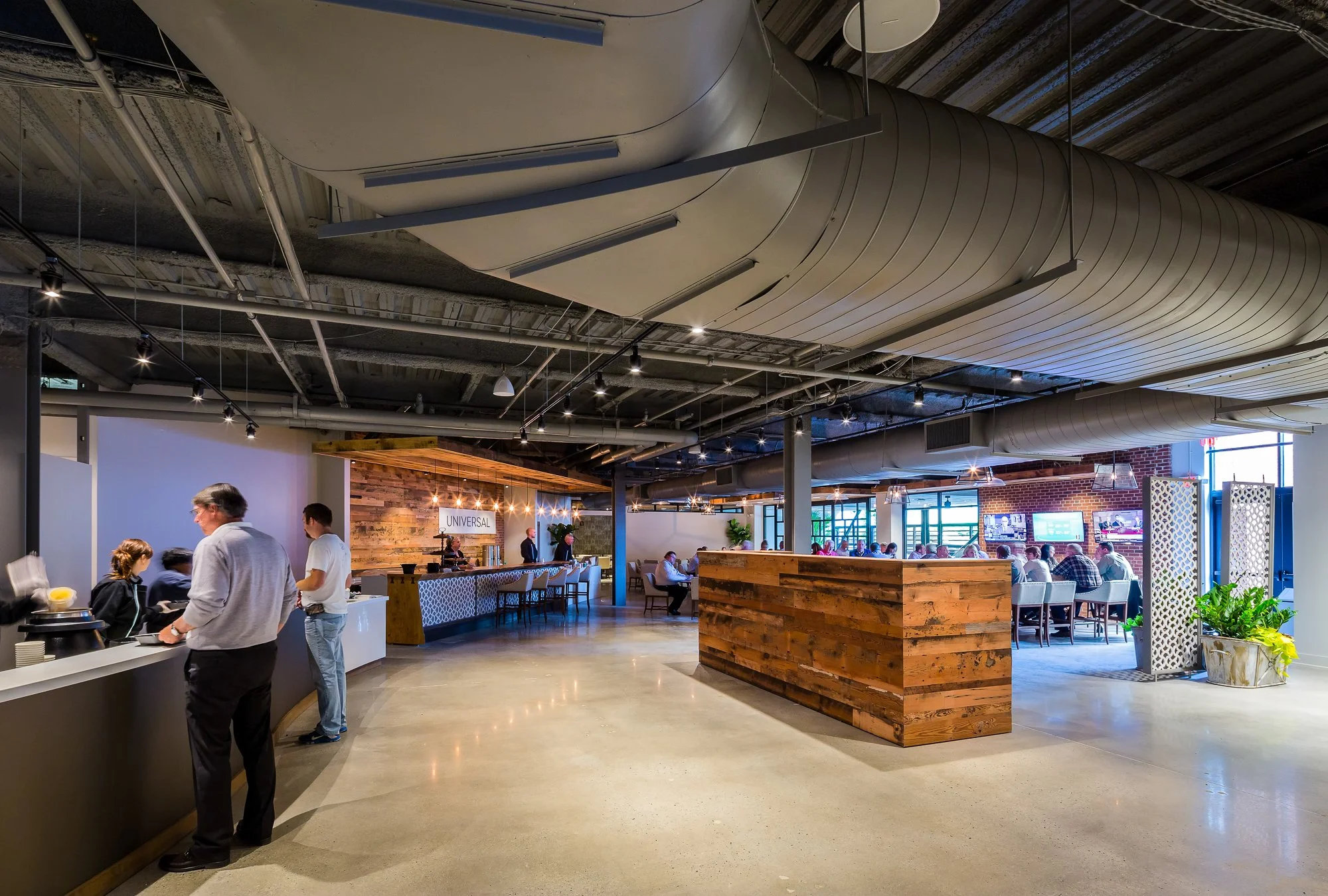 Interior of a modern restaurant with exposed ductwork, track lighting, and a mix of wooden and brick decor. Tables are occupied by customers, and there is a counter with staff serving food.
