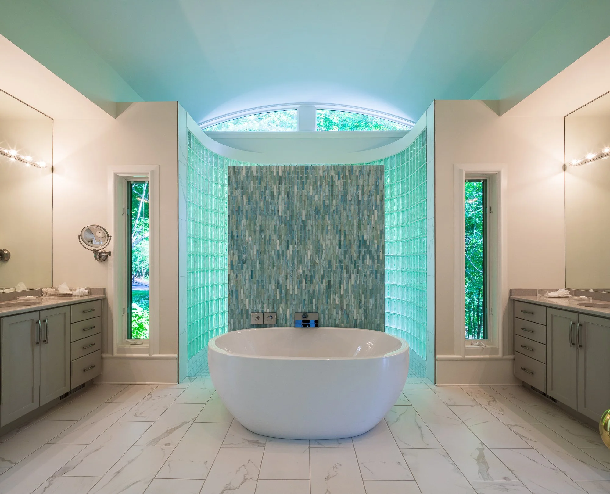 Luxurious bathroom with white marble tile flooring, a freestanding bathtub, large windows with greenery outside, gray cabinetry, and glass block walls.