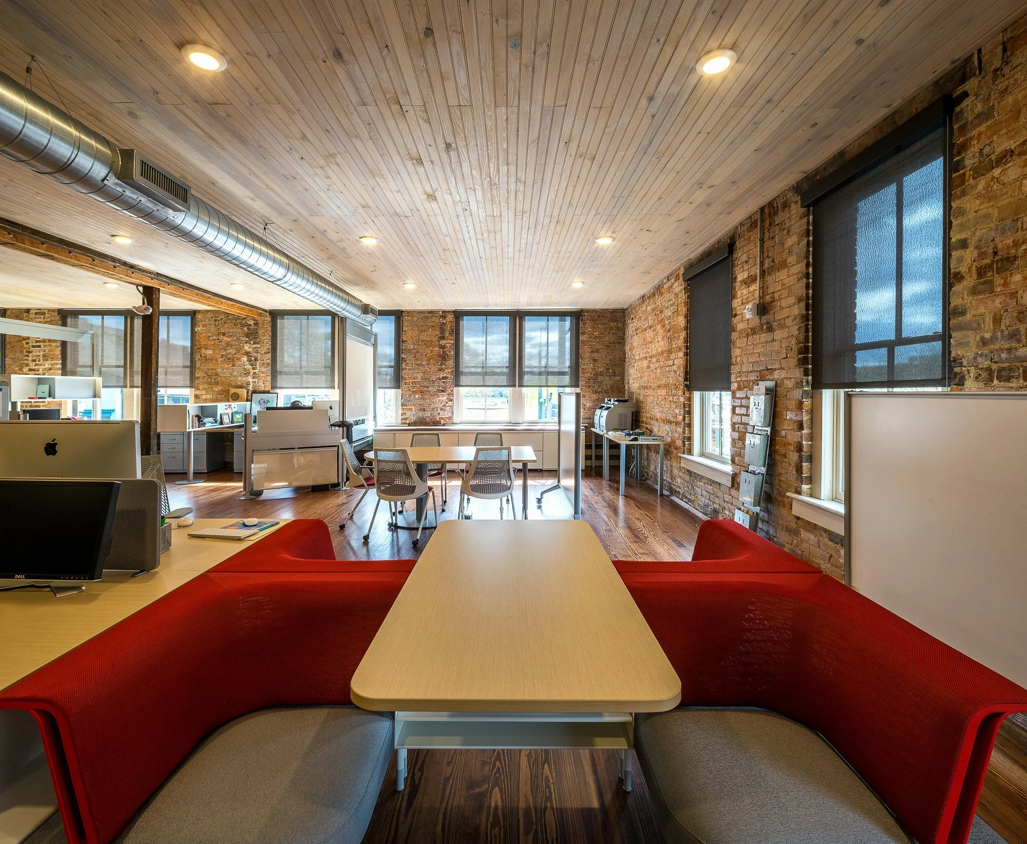 Modern office space with exposed brick walls, wooden ceilings, and large windows with black shades. Office furniture includes tables, chairs, and computer desks.