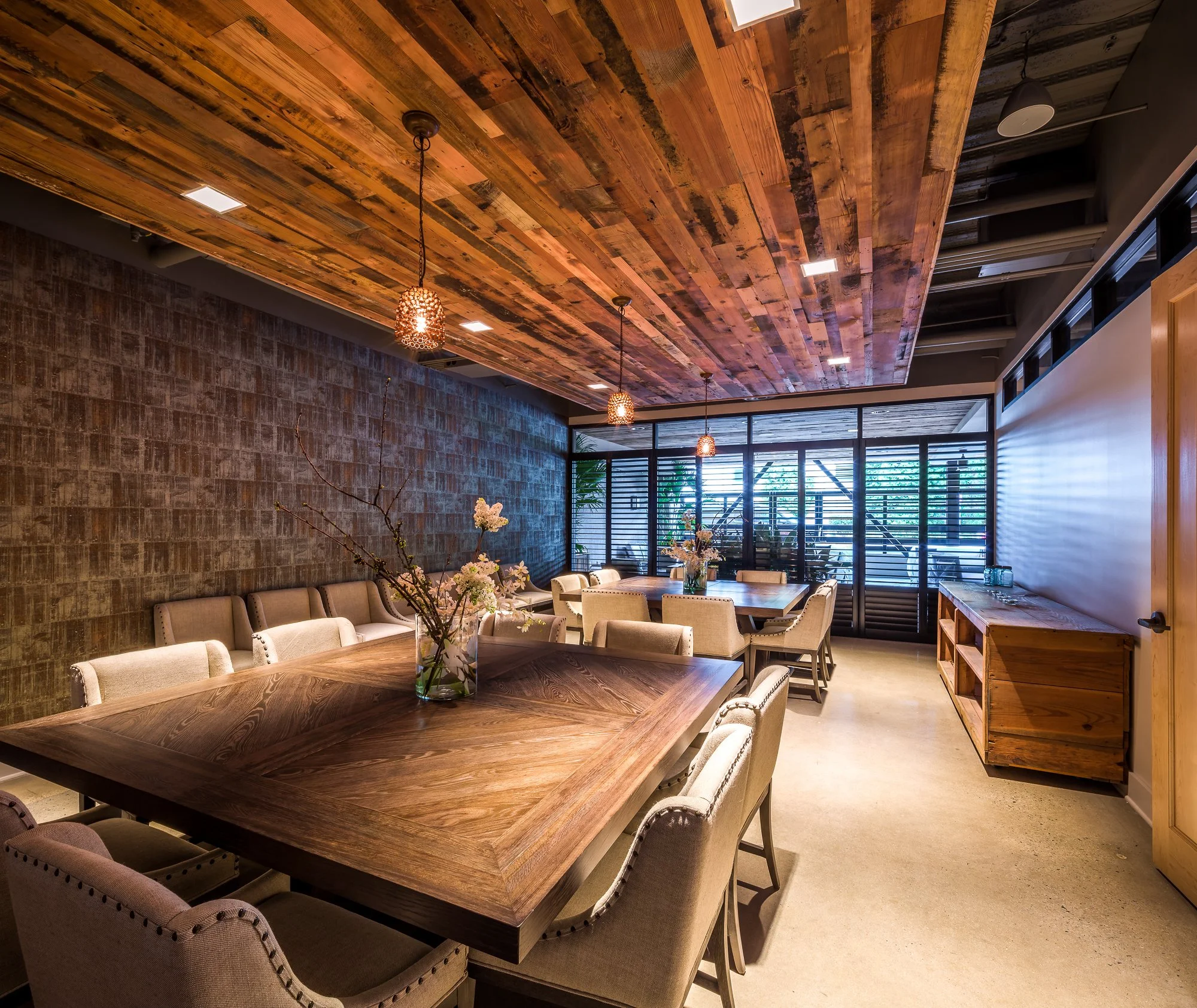 A dining area with a large wooden table, beige upholstered chairs, a glass vase with white flowers, wooden ceiling with hanging pendant lights, brick wall, large windows with black shutters, and greenery outside.