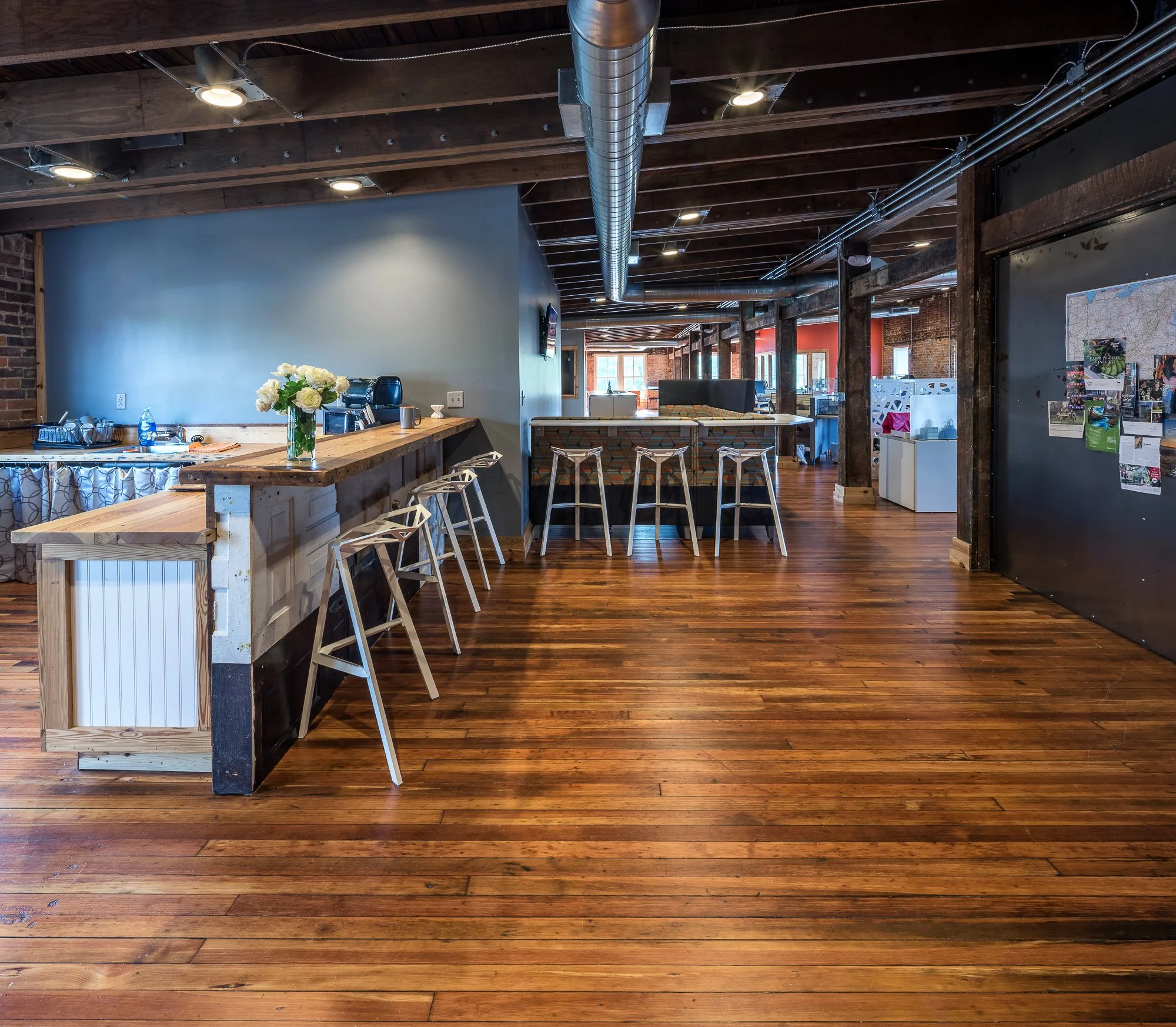 Interior of an industrial-style cafe or coworking space with wooden floors, exposed brick walls, a long wooden bar with white and wooden chairs, modern lighting, and large windows in the background.