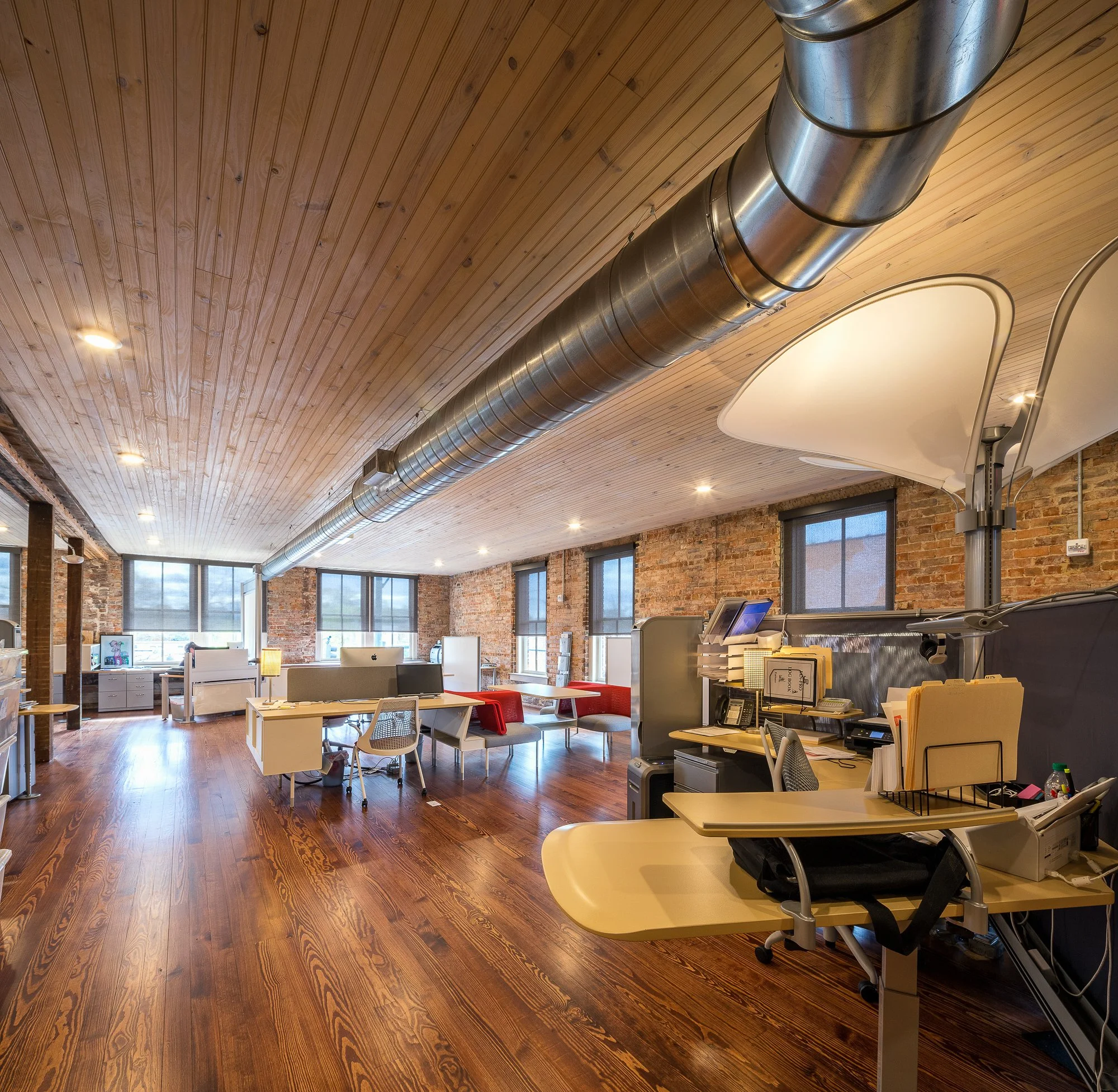 Open office space with wooden ceiling, large ventilation duct, brick walls, desks, chairs, and windows.