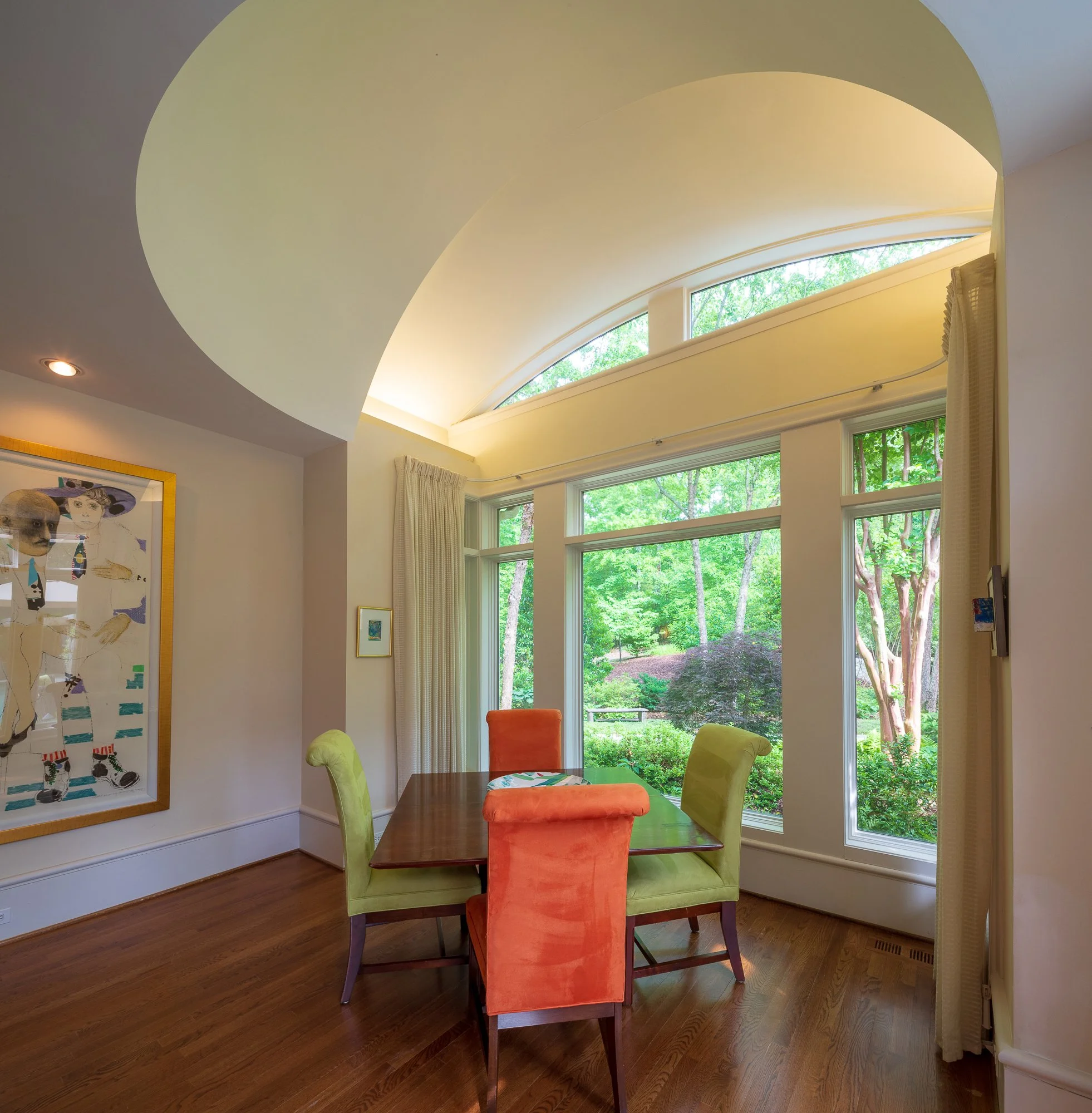 A dining room with a wooden table and four colorful chairs, two green, one orange, and one yellow. Large windows showing green trees outside, with curtains and framed artwork on the walls. Ceiling lights are also visible.
