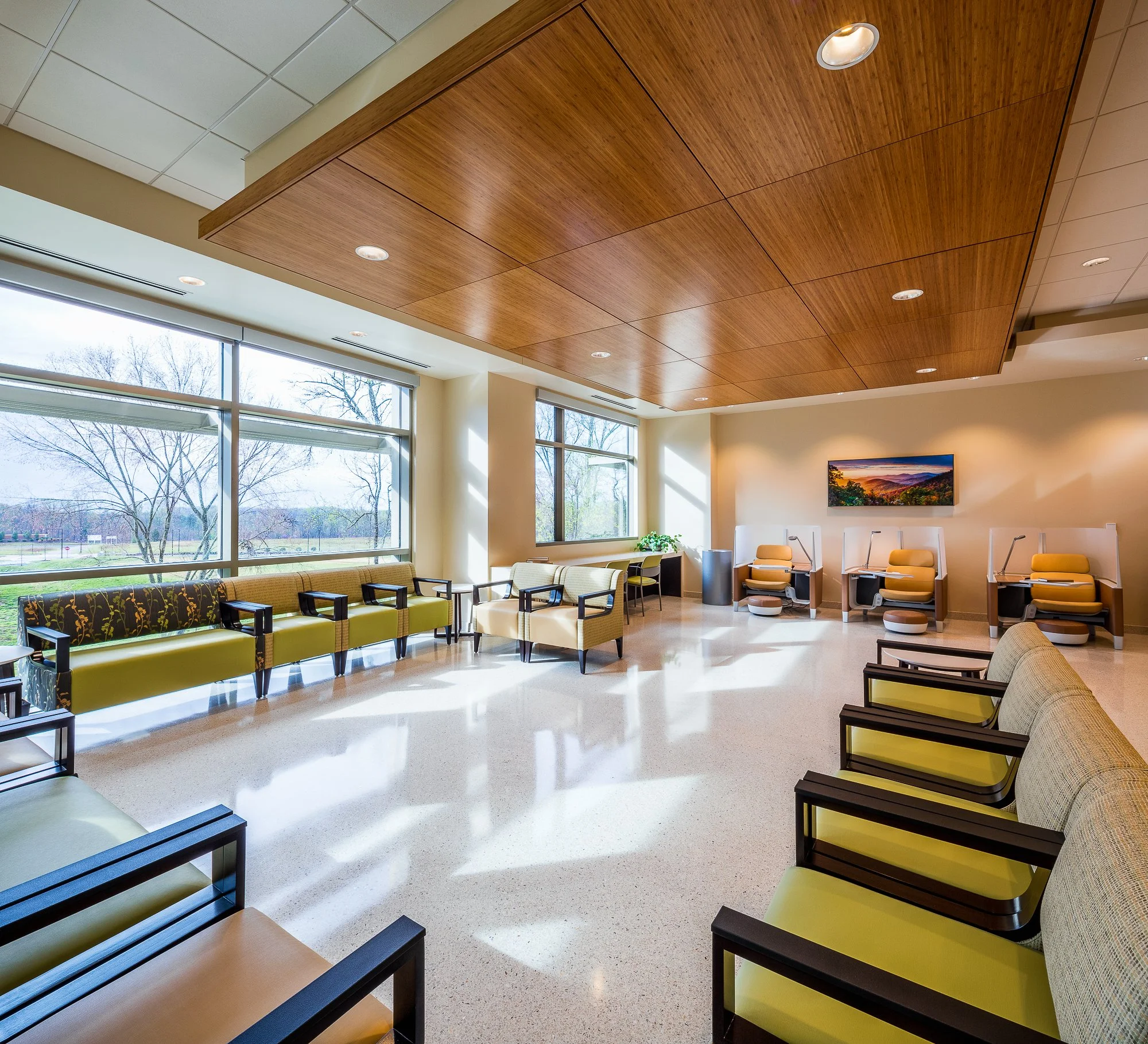 Interior view of a waiting room or lounge area with large windows, modern chairs, a mounted flat-screen TV, and a wooden ceiling with embedded lights.