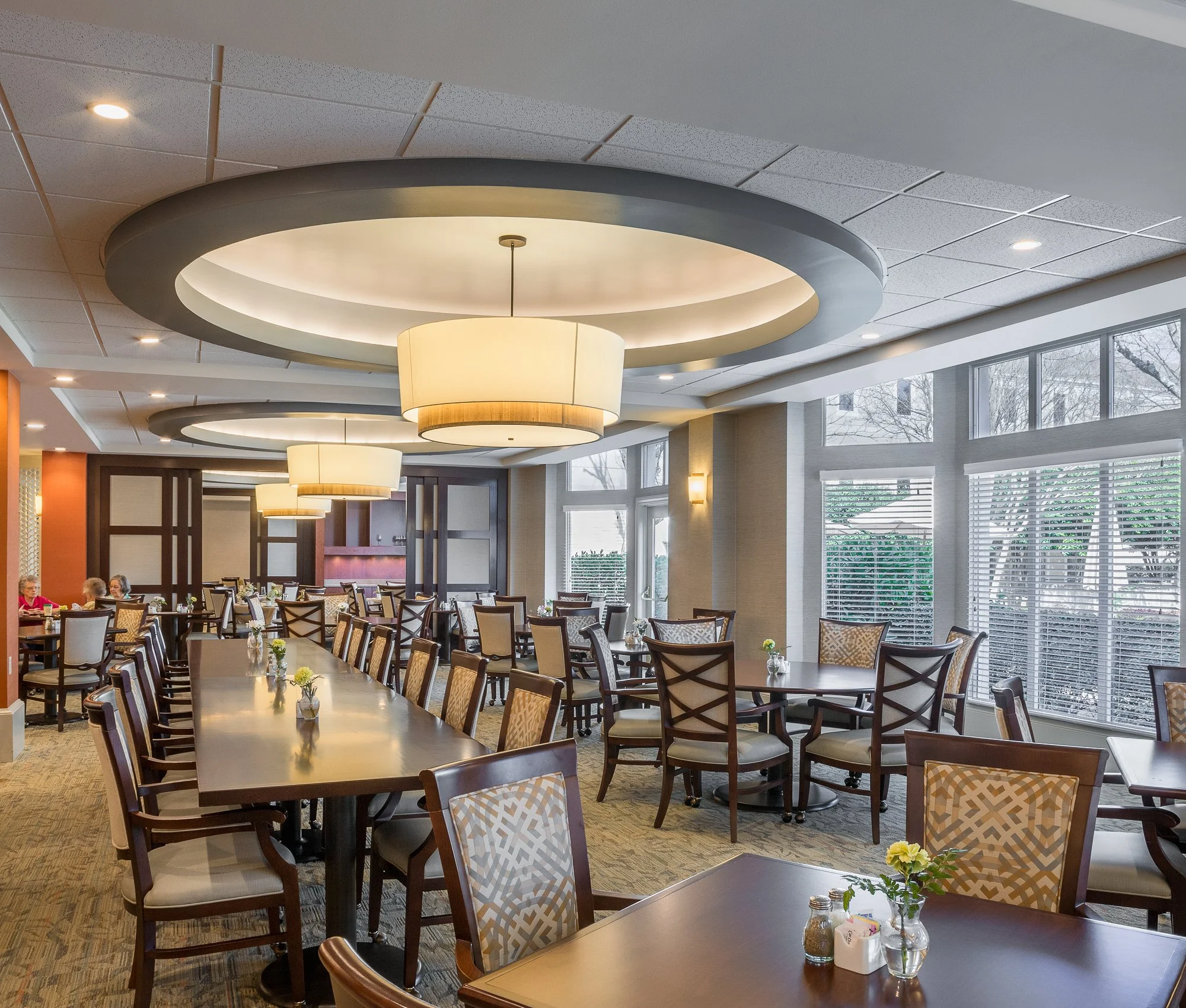Casual restaurant dining area with round and rectangular tables, wooden chairs with patterned cushions, large windows, modern ceiling lights, and some elderly patrons seated at tables.