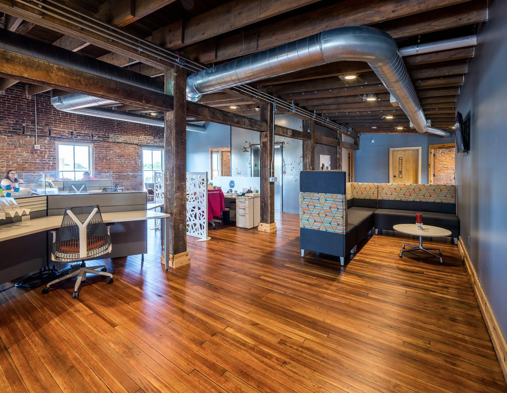 Open office space with exposed brick walls, wooden flooring, and visible ceiling beams with ductwork. There are desks, chairs, and a seating area with patterned upholstery.