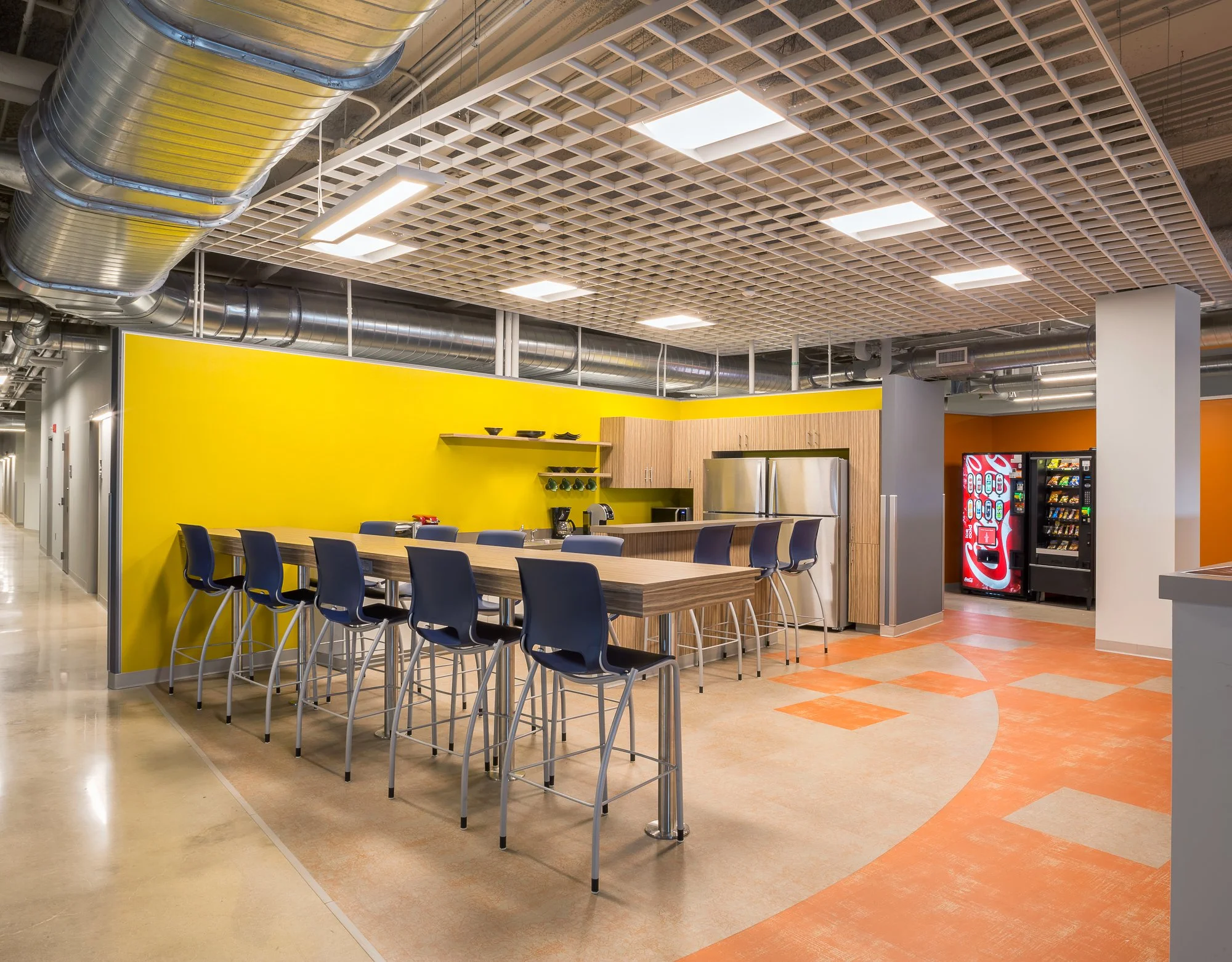 A cafeteria with a long wooden table and blue chairs, yellow wall, and exposed industrial ductwork ceiling with bright rectangular lights.