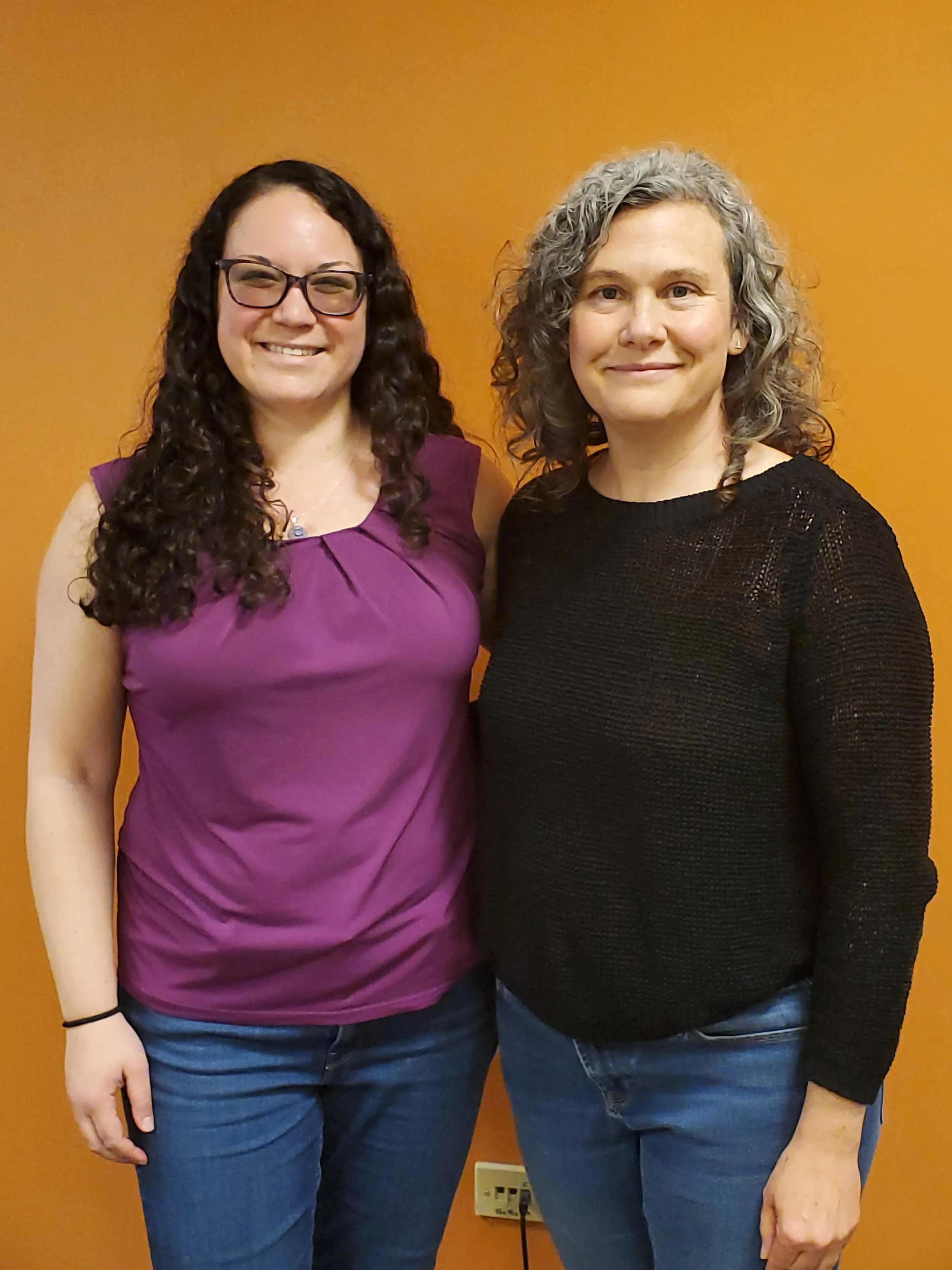 Two women standing side by side in front of an orange wall, smiling at the camera.