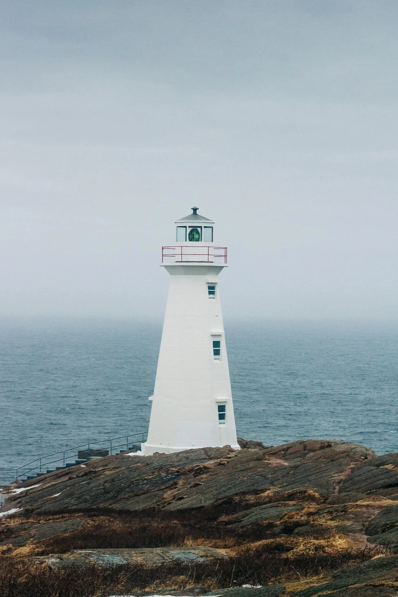 A white lighthouse on a rocky shoreline facing the ocean under a cloudy sky.