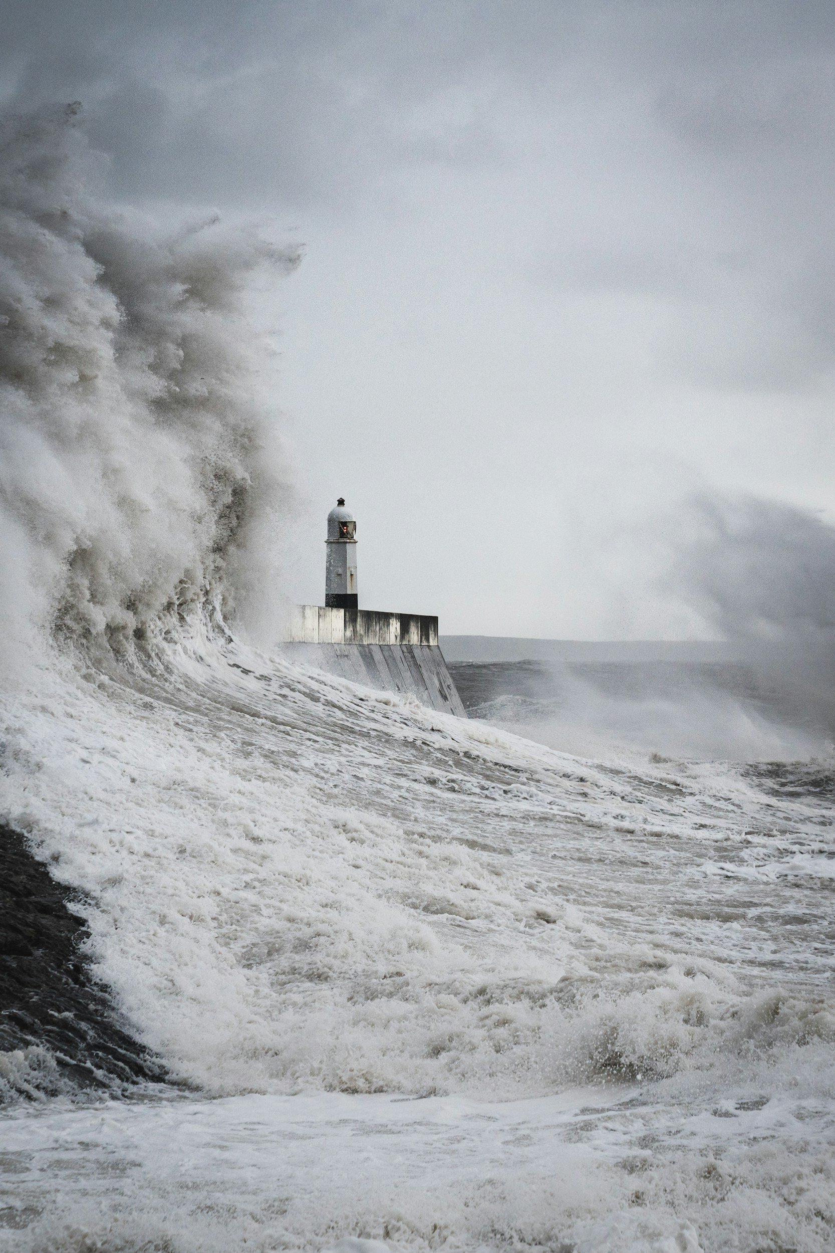 A lighthouse near the coast with large waves crashing against the seawall during stormy weather.