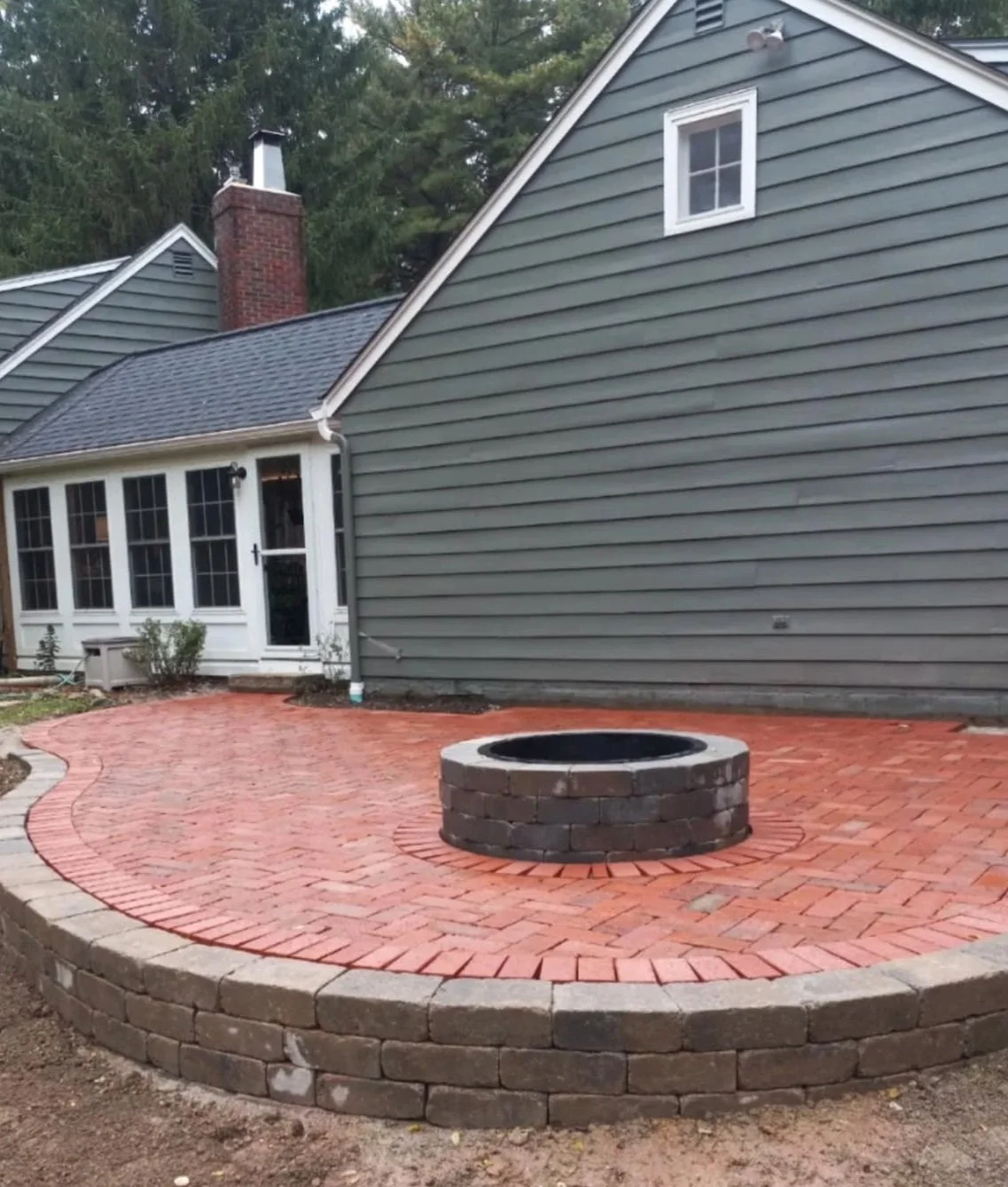 A backyard patio with red brick pavers, featuring a circular fire pit made of stacked bricks, close to the house with gray siding. There are plants near the house and a small window on the side.