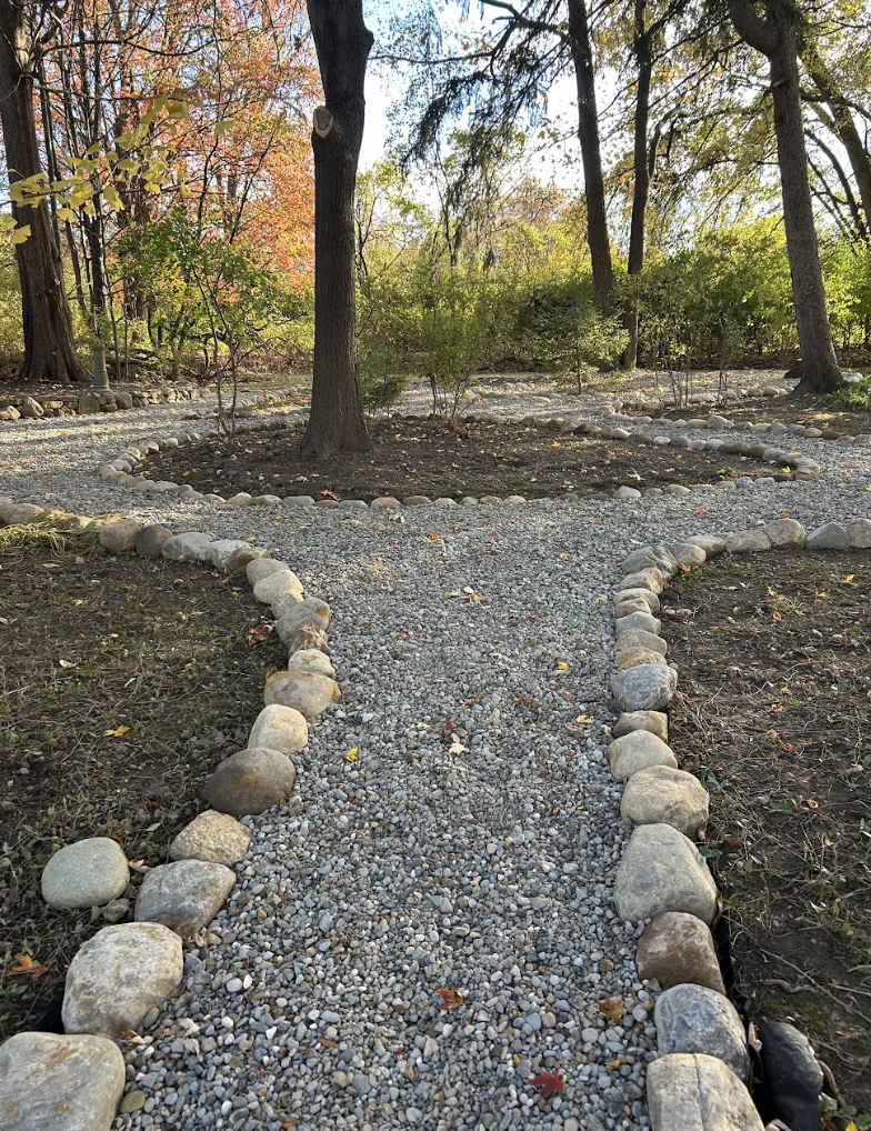 A gravel pathway lined with rocks winds through a wooded area with trees that have green, yellow, and red leaves.