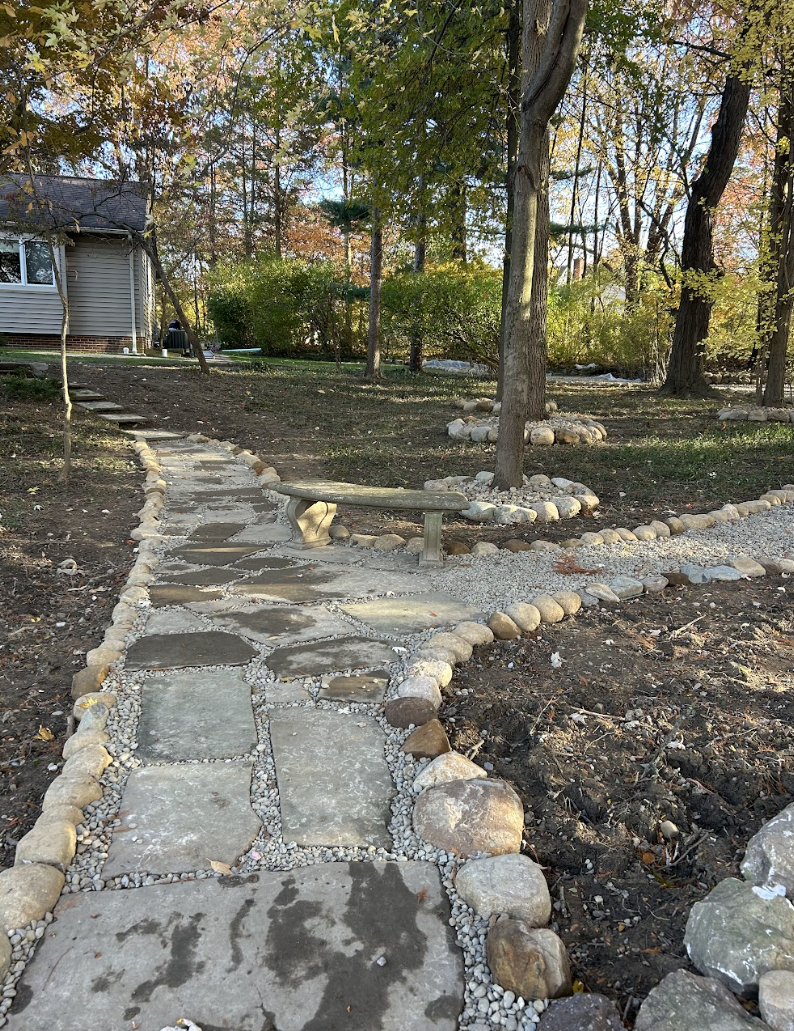 A stone pathway in a backyard with a wooden bench, trees, and a house in the background.