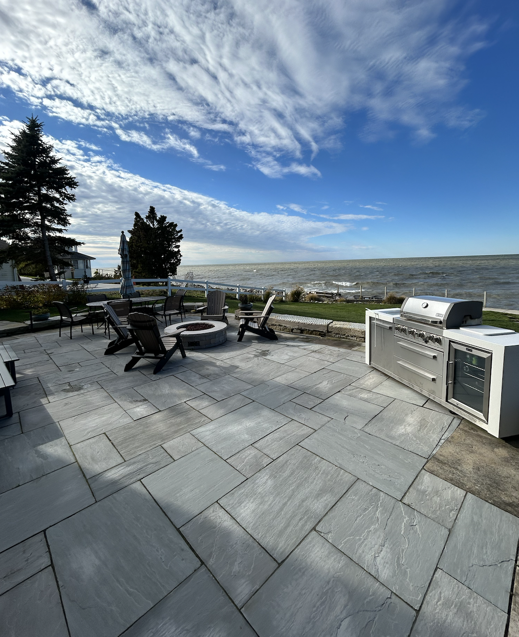 A spacious backyard patio overlooking a body of water with outdoor furniture, a fire pit, and a built-in grill, under a partly cloudy sky.