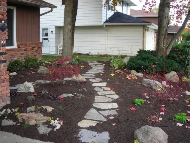 A garden path made of irregular flat stones winding through a garden with various plants and trees, with a house in the background.