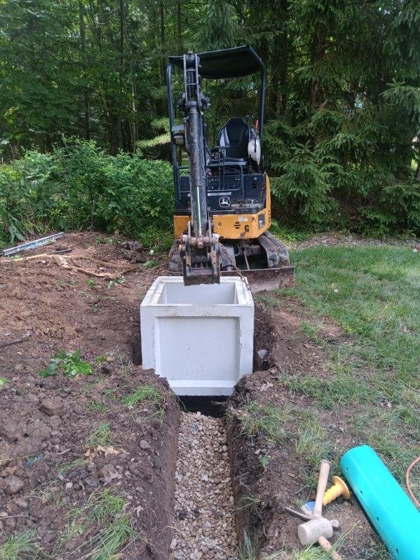 A small excavator placing a large concrete utility box into a trench during a construction or landscaping project in a backyard.