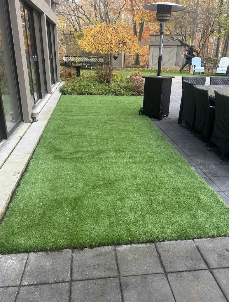 A backyard patio with artificial green grass, outdoor chairs, a heater, and trees with autumn foliage in the background.