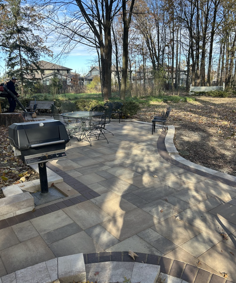 An outdoor patio with paved stone flooring, a barbecue grill, metal patio chairs around a round table, and trees in the background. A person is seen in the upper left corner near a bench.
