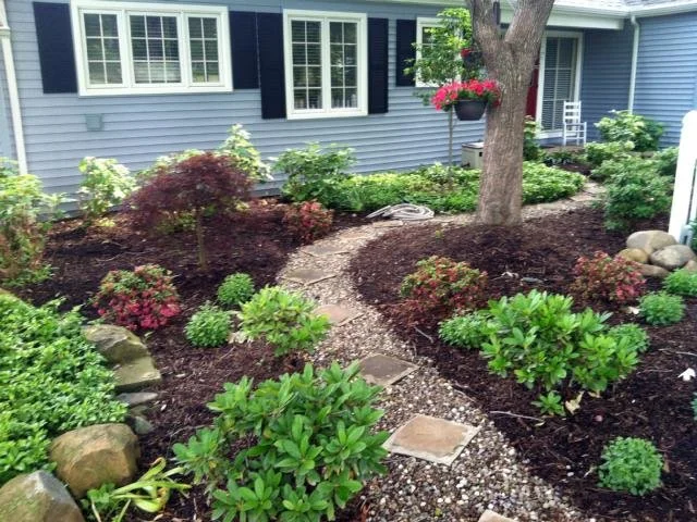 A garden with a winding pathway made of small stones, surrounded by various green and flowering shrubs, and a tall tree with a hanging flower basket nearby. There is a house with blue siding and white trim in the background.