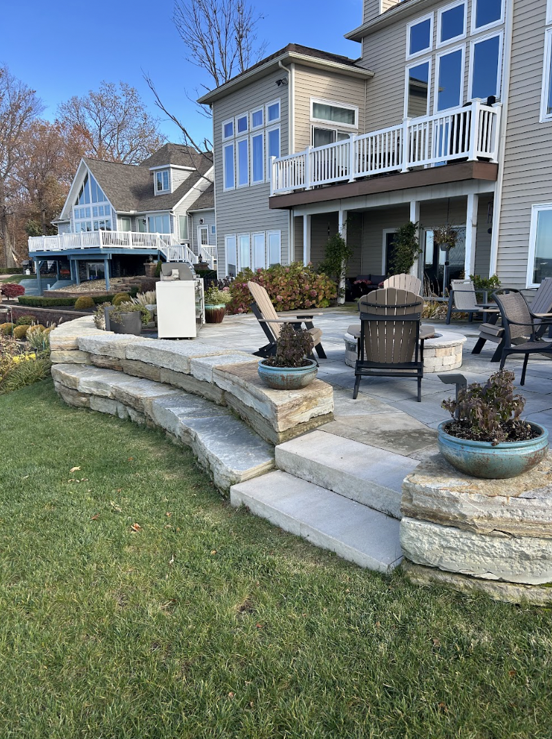 A backyard patio with Adirondack and other outdoor chairs, surrounded by potted plants, stone walls, and a house with a balcony.