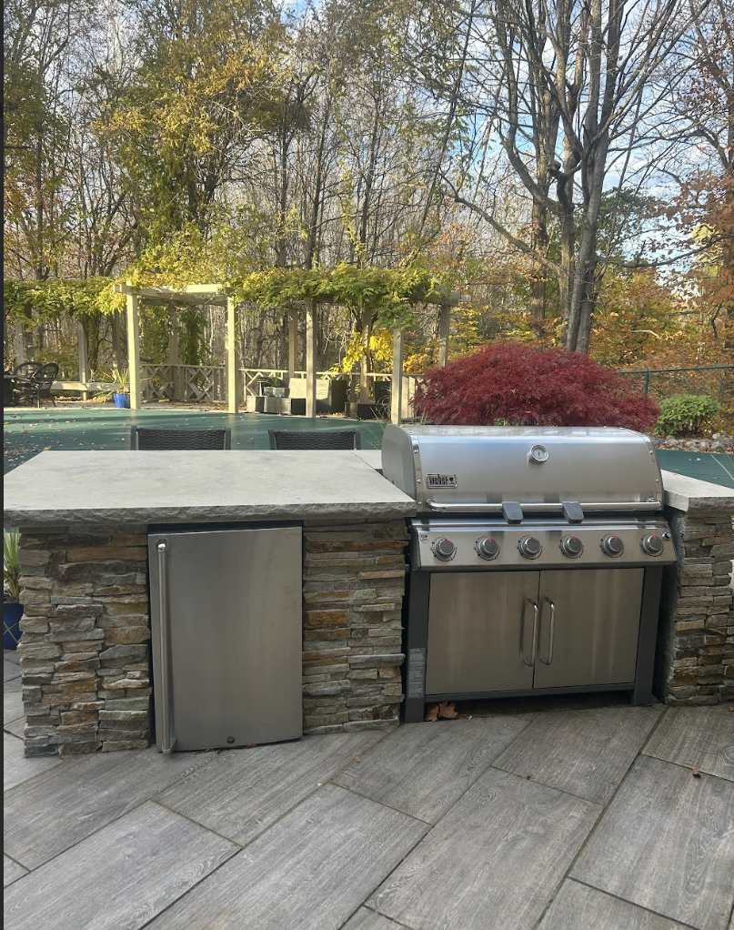Outdoor kitchen with a stainless steel grill, stone counter, small fridge, and surrounding trees and backyard landscape.
