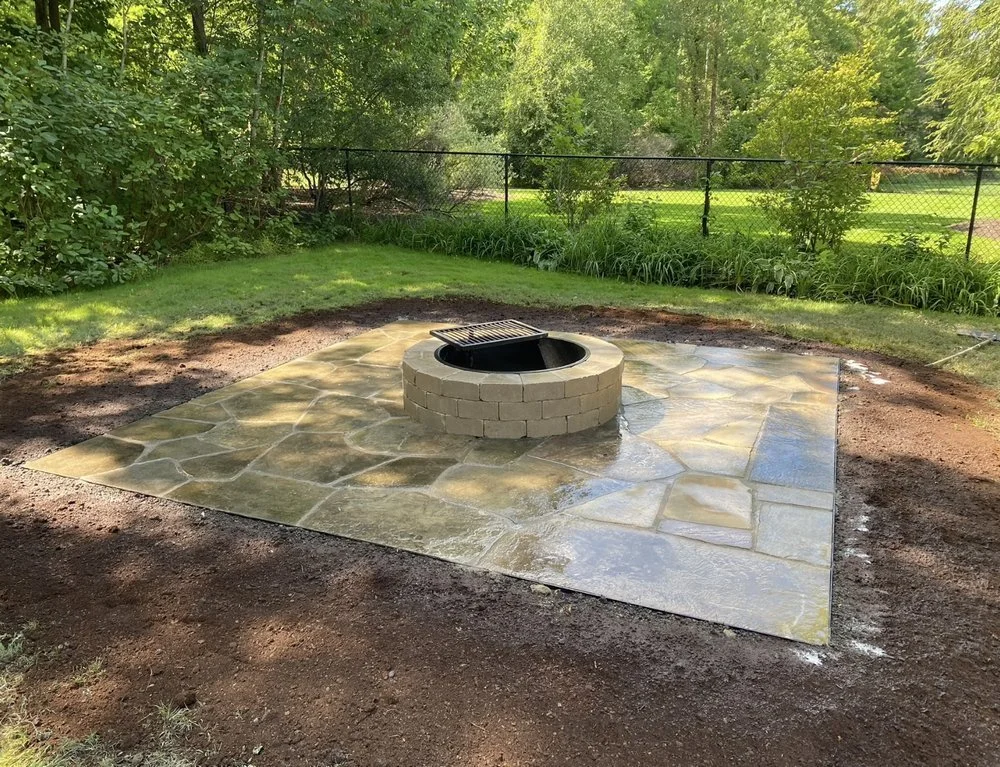 A newly built fire pit with a black center and a metal grate, surrounded by stone pavers on a grassy backyard.