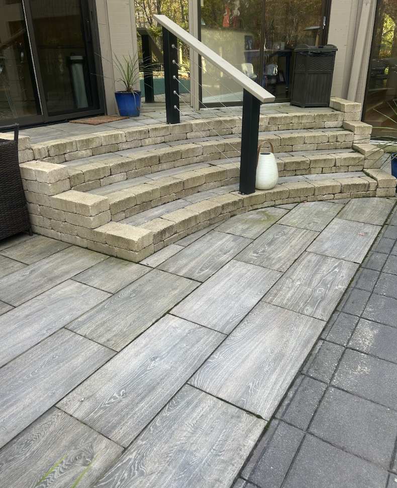 Concrete steps with black railing leading to a glass door entrance, surrounded by potted plants and outdoor furniture, with a wood and concrete patio.