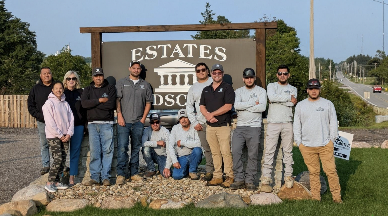 Group of ten people, the Estates Landscaping team, standing in front of a sign that reads 'ESTATES LANDSCAPING' with the company's logo, outdoors on a sunny day with trees and a road in the background.