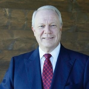 A smiling older man with gray hair, dressed in a navy suit, white shirt, and patterned red tie, standing in front of a stone wall.