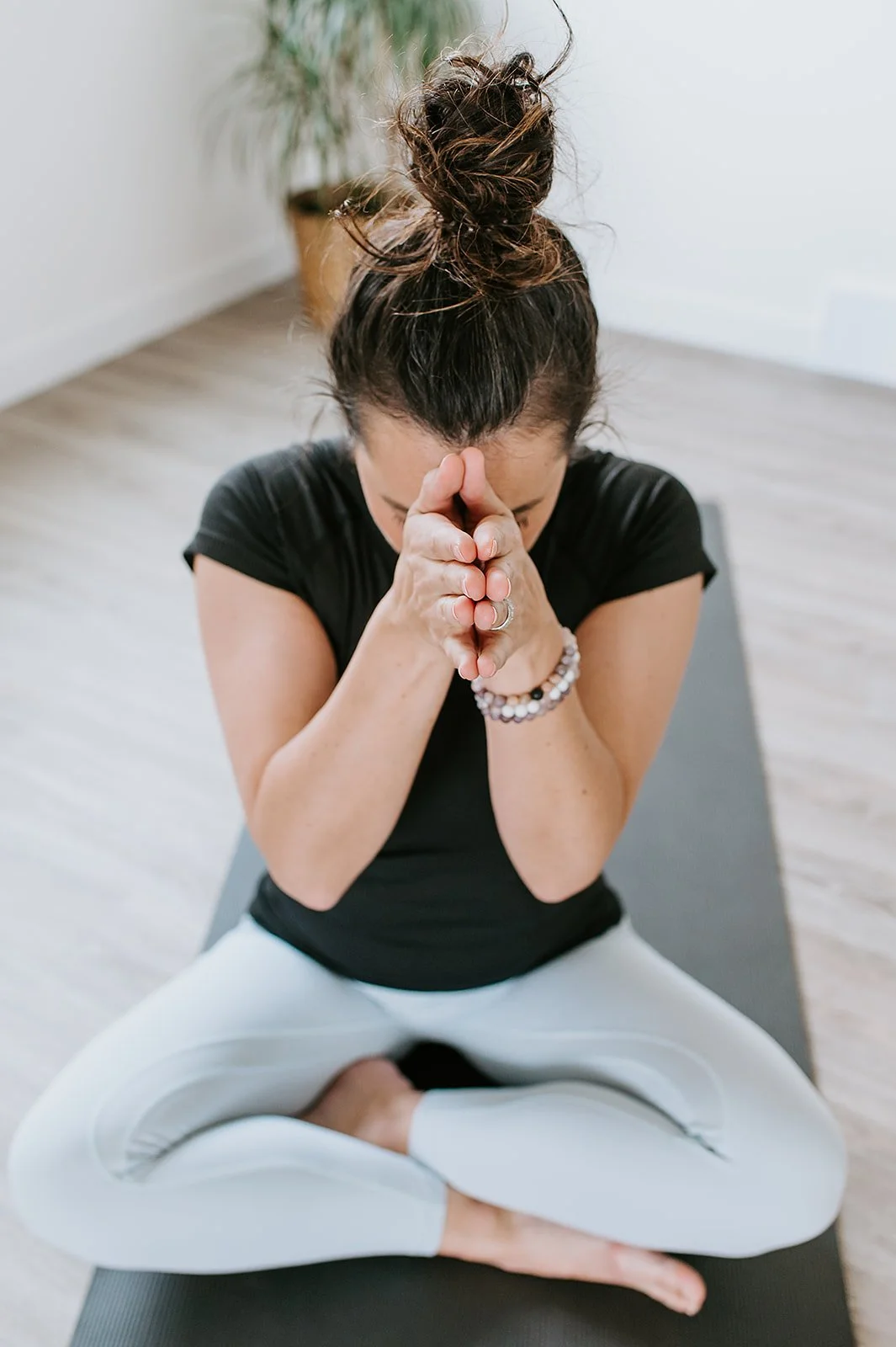 Woman practicing yoga, sitting cross-legged on a mat with hands pressed together in prayer position, head bowed, in a bright room.