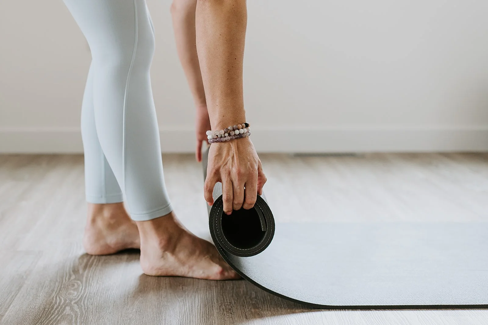 Person in white leggings unrolling a black yoga mat on a wooden floor.