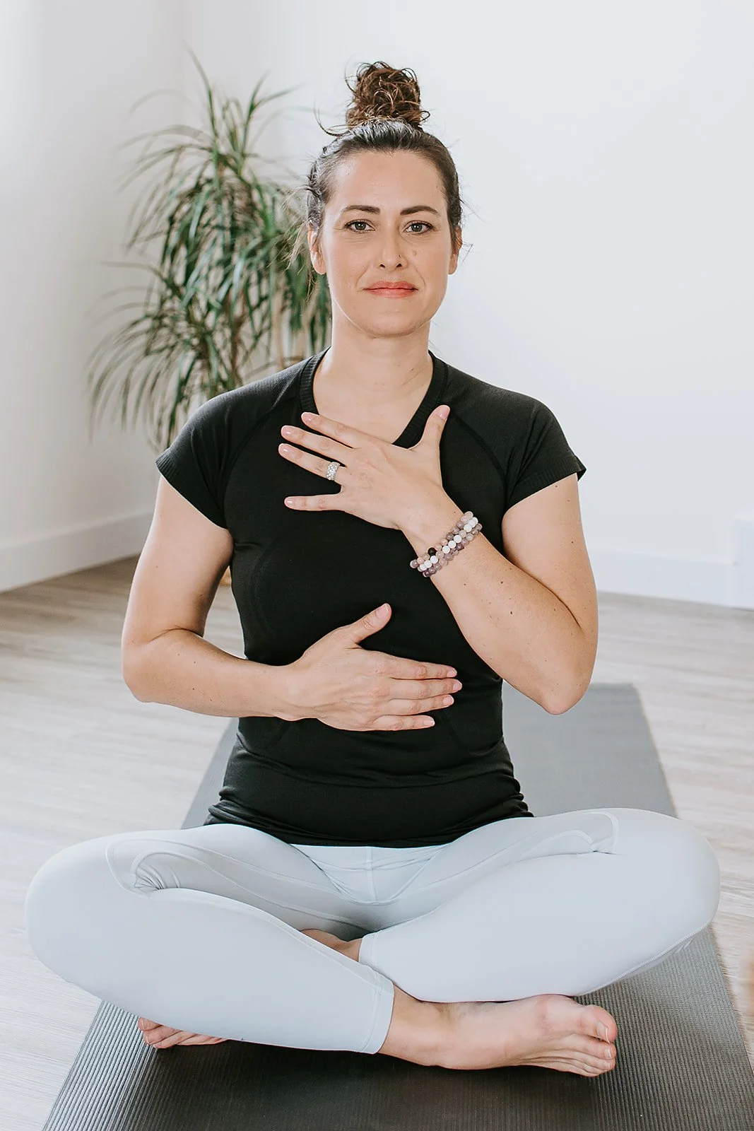 A woman practicing yoga in a seated position on a yoga mat, with one hand on her chest and the other on her abdomen, indoors with a plant in the background.