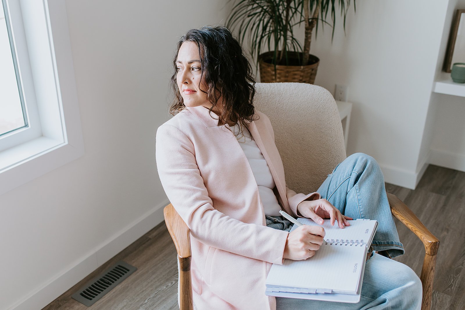 Woman sitting in a chair near a window holding a notebook and pen, looking thoughtfully outside, with a plant and shelf in the background.