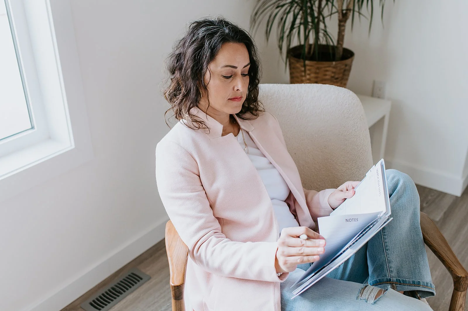 Woman with dark curly hair wearing a light pink blazer sitting on a beige chair reading a notebook with notes.